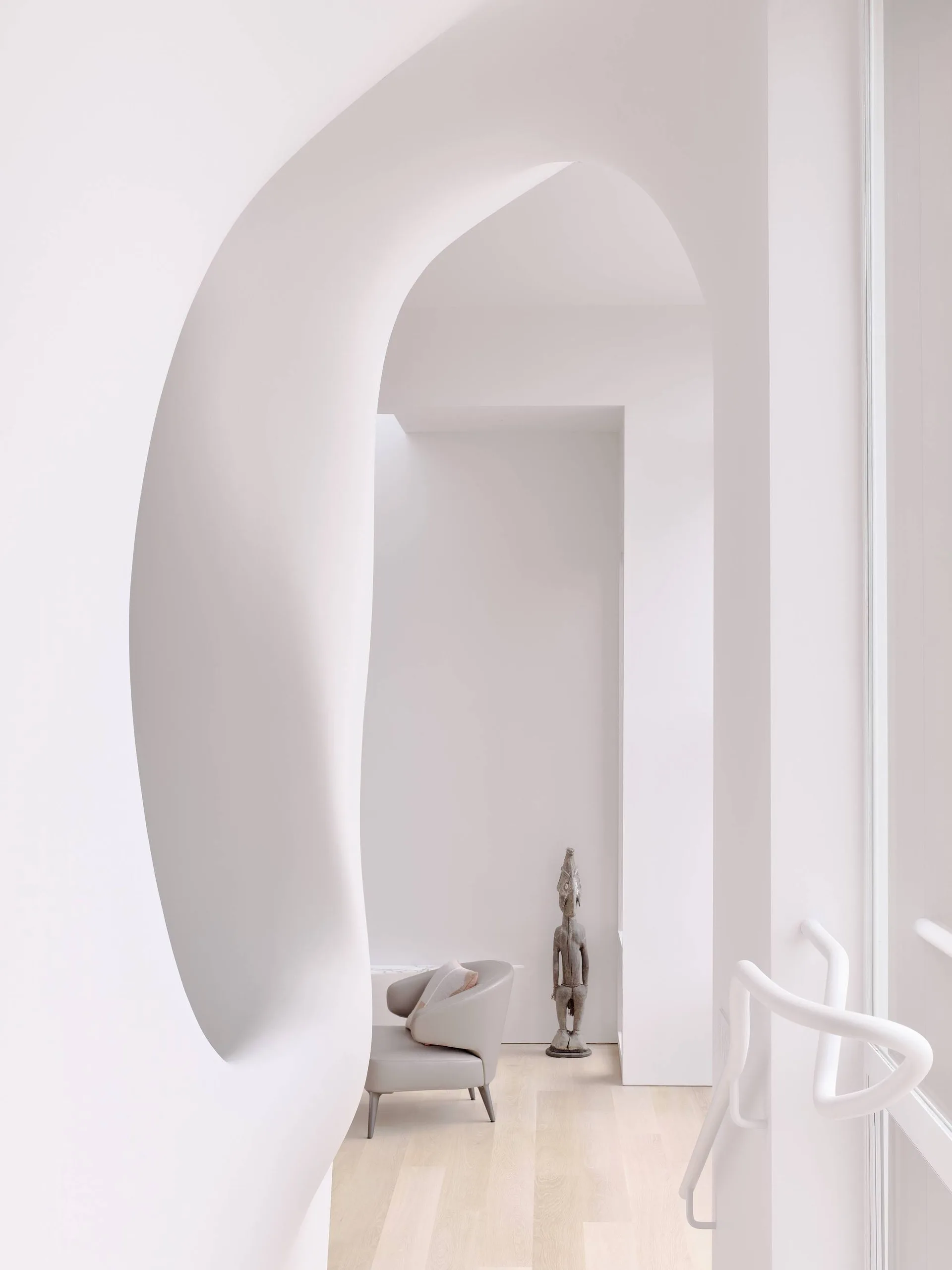 A minimalist white hallway features a large curved plaster archway framing a grey armchair and a tall wooden sculpture on light wood floors.
