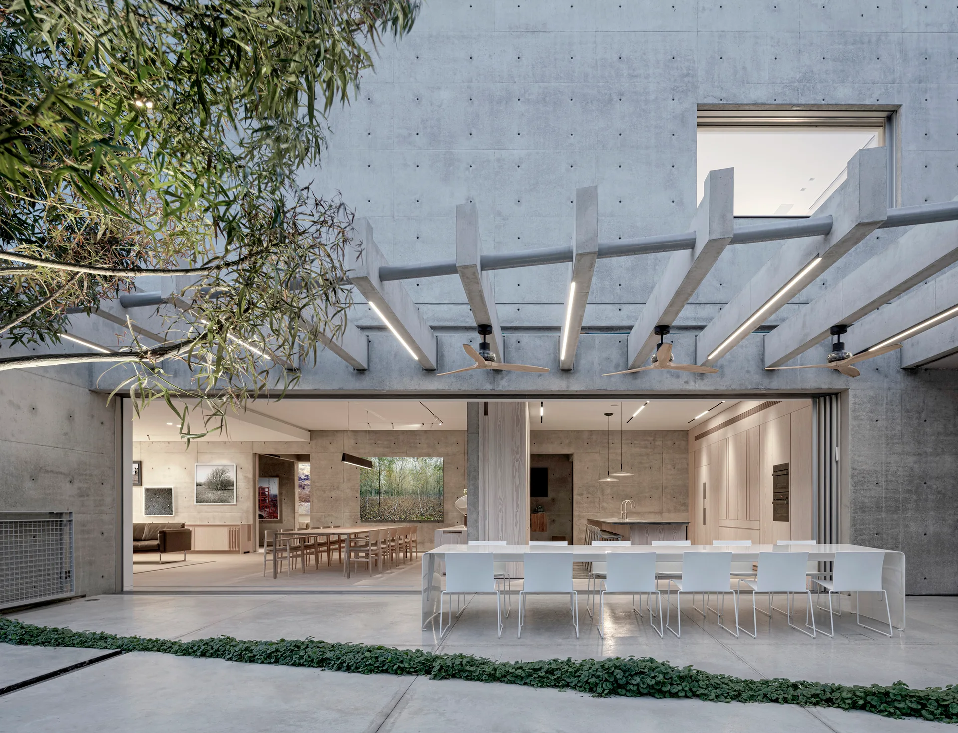 A long white dining table stands on a concrete patio beneath a concrete pergola with integrated lights, next to sliding glass doors.