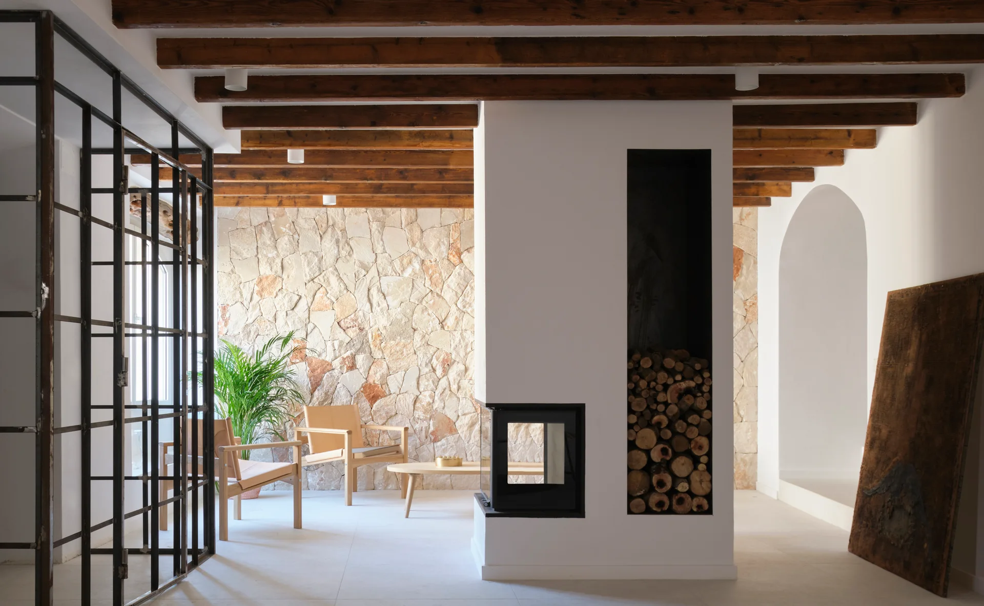 A modern living space featuring a central white fireplace column with firewood storage, exposed ceiling beams, and a rough stone accent wall.