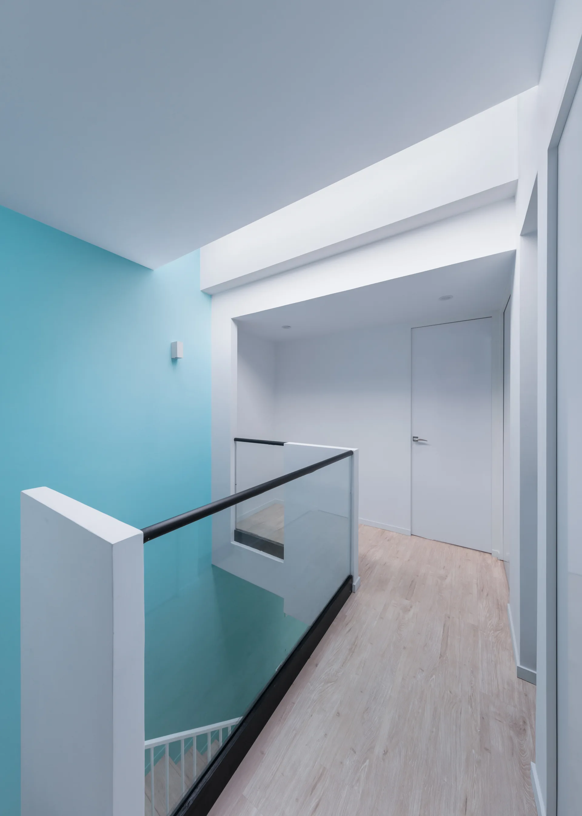 A contemporary hallway landing featuring light wood floors, a glass staircase railing, and a vibrant blue accent wall under natural overhead light.