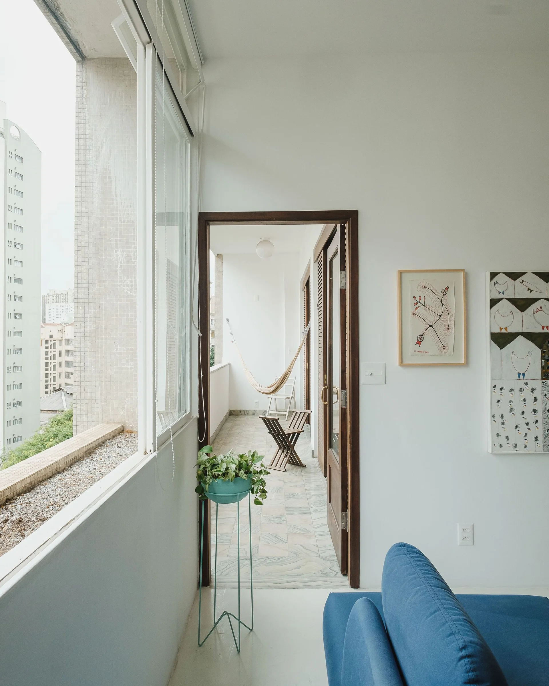 A bright white living room with a blue sofa features an open wood-framed doorway leading to a narrow balcony with a hammock and folding chairs.