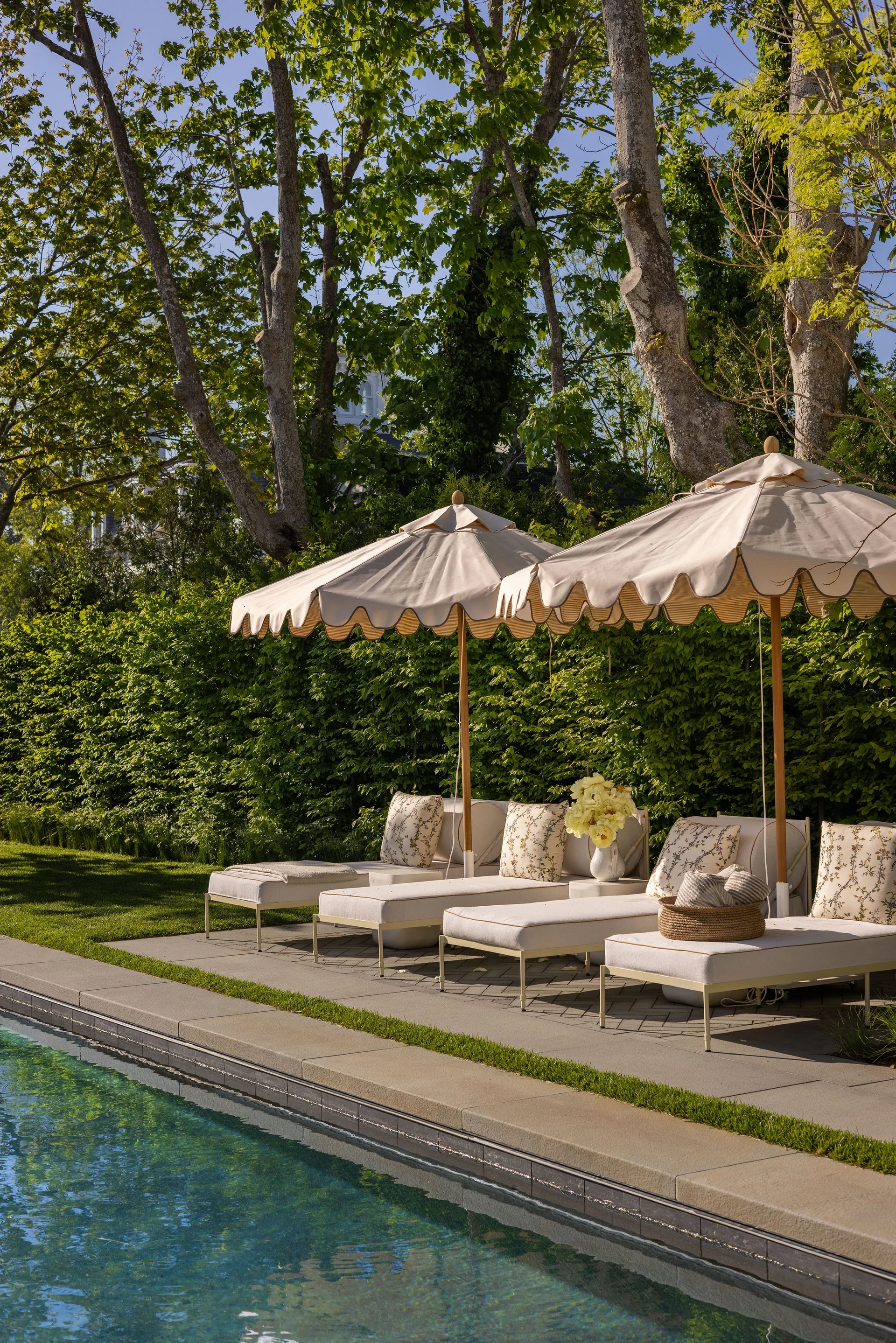 Four off-white chaise loungers sit beside a swimming pool under two scalloped umbrellas, bordered by a dense green hedge.