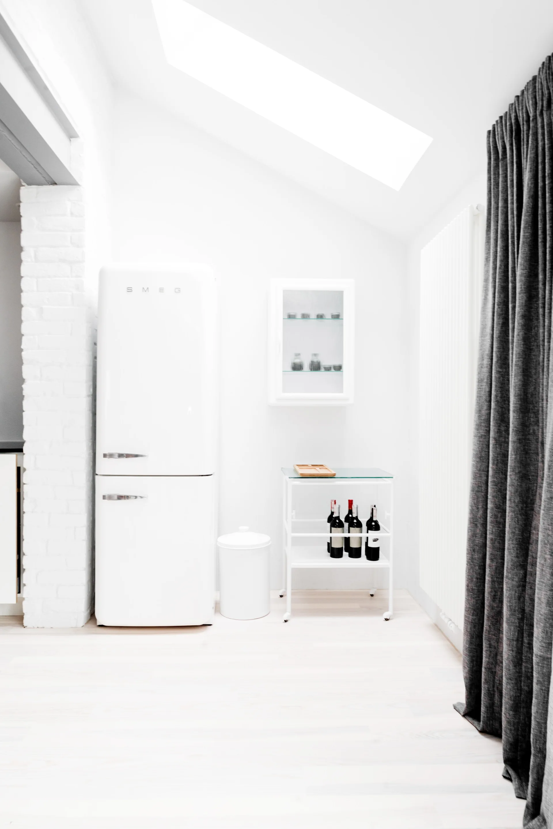 A minimalist white kitchen corner featuring a retro white refrigerator, a rolling bar cart with wine, and a white brick accent wall section.