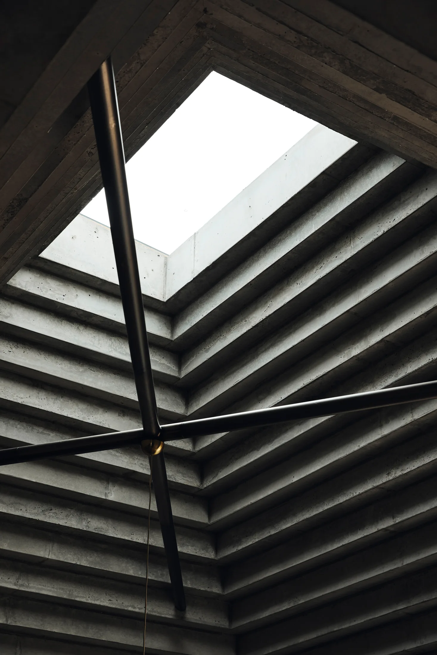 A low-angle view of a stepped grey concrete ceiling with a rectangular skylight and intersecting black metal support beams.