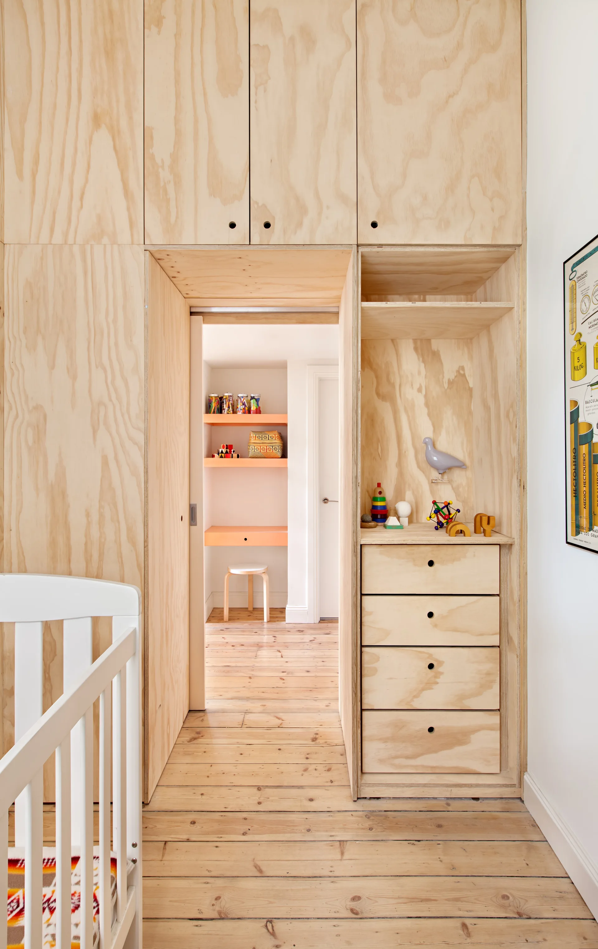 A light wood plywood storage wall surrounds a doorway leading to a desk area in a bright nursery with a white crib in the foreground.