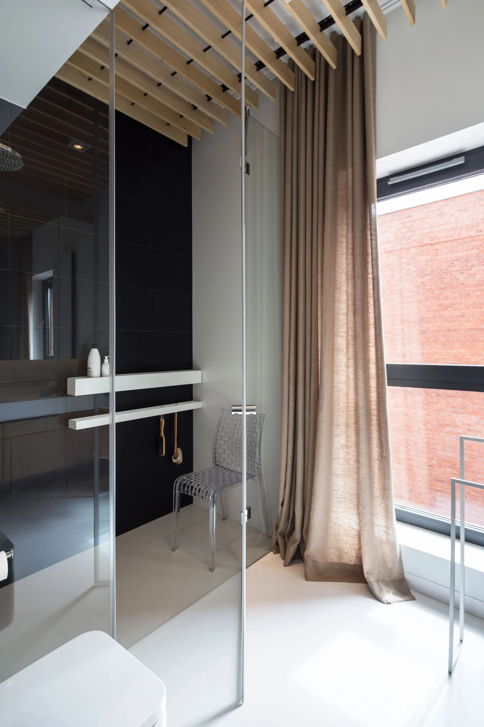 A modern bathroom features a glass partition, a transparent plastic chair, a slatted wood ceiling, and floor-length beige curtains.