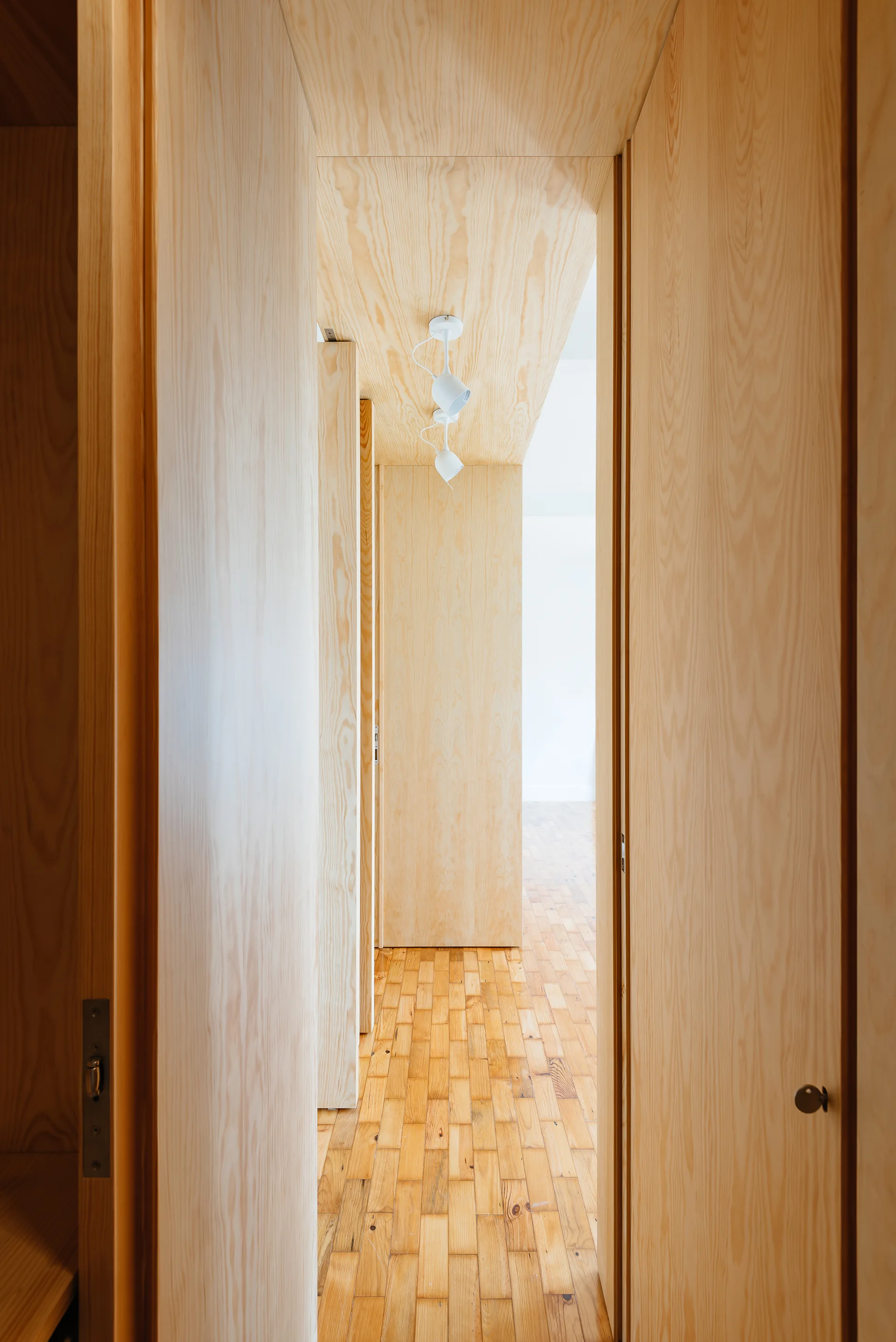 An interior hallway features light plywood panels on the walls and ceiling, flush wooden doors, and a natural parquet block floor.