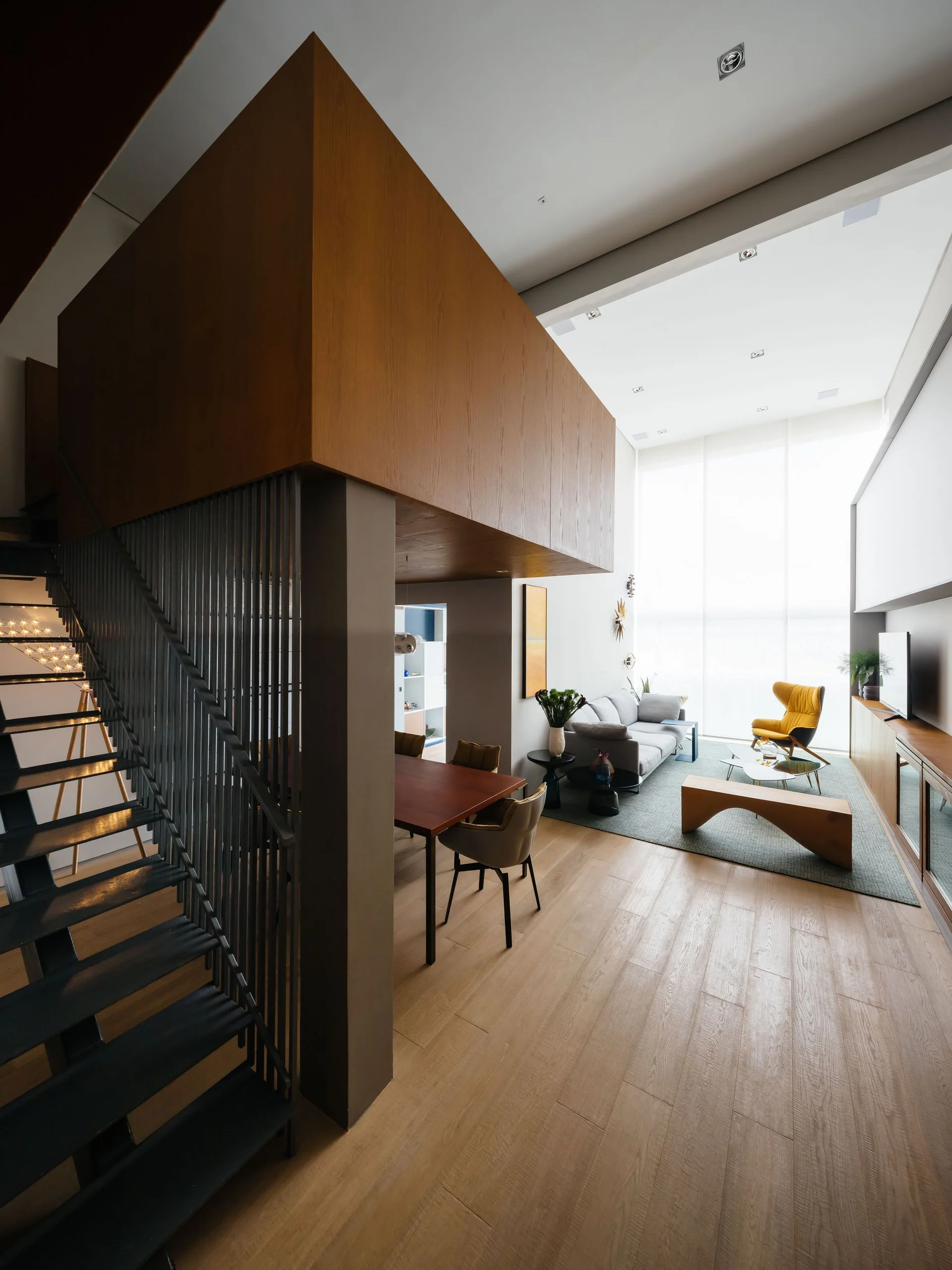 A modern open-plan living room features a wooden mezzanine structure, a metal staircase, a gray sofa, and a yellow armchair under a high ceiling.