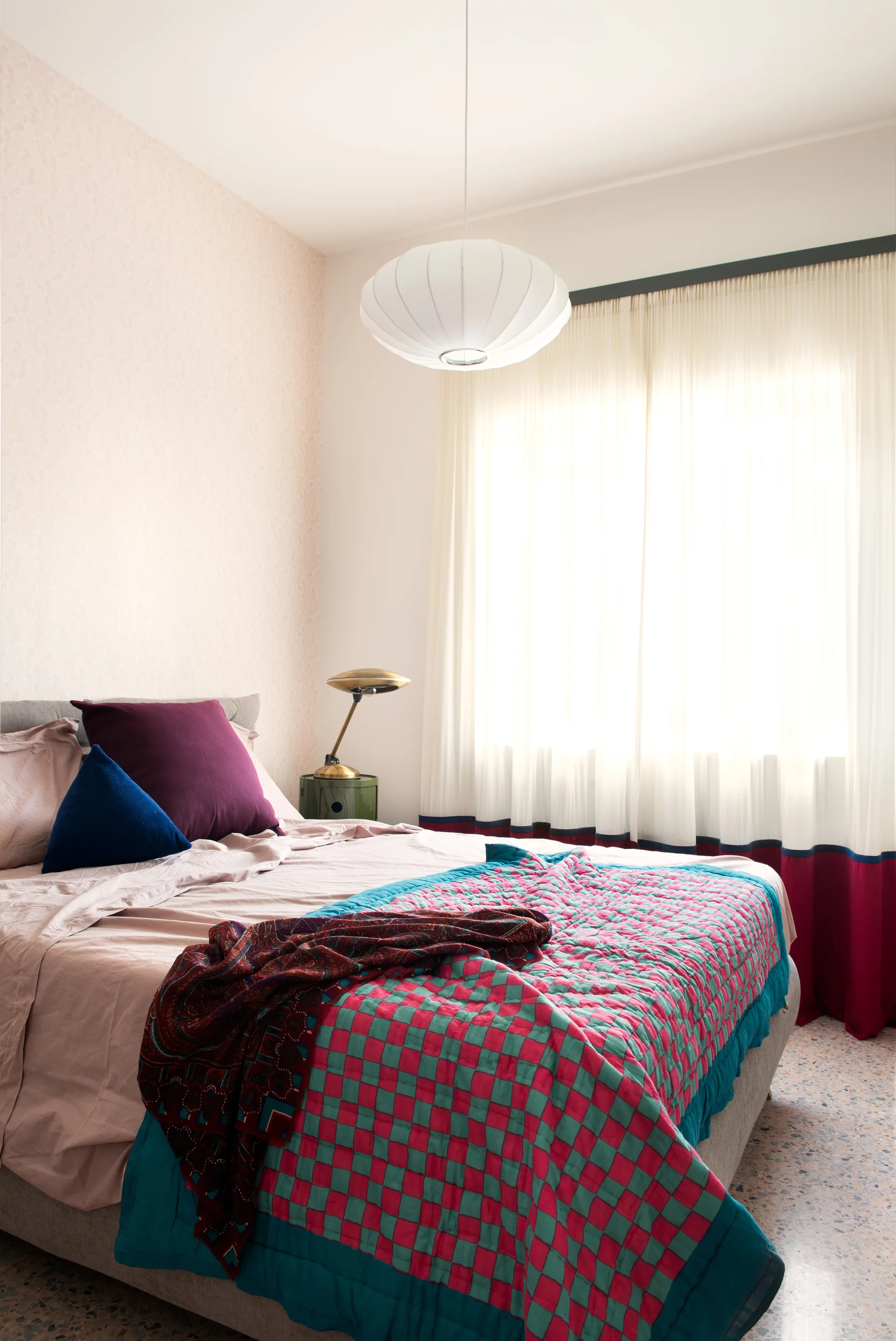 An eclectic bedroom featuring an upholstered bed with a pink and teal checkered quilt, faint pink floral wallpaper, and a paper pendant light.
