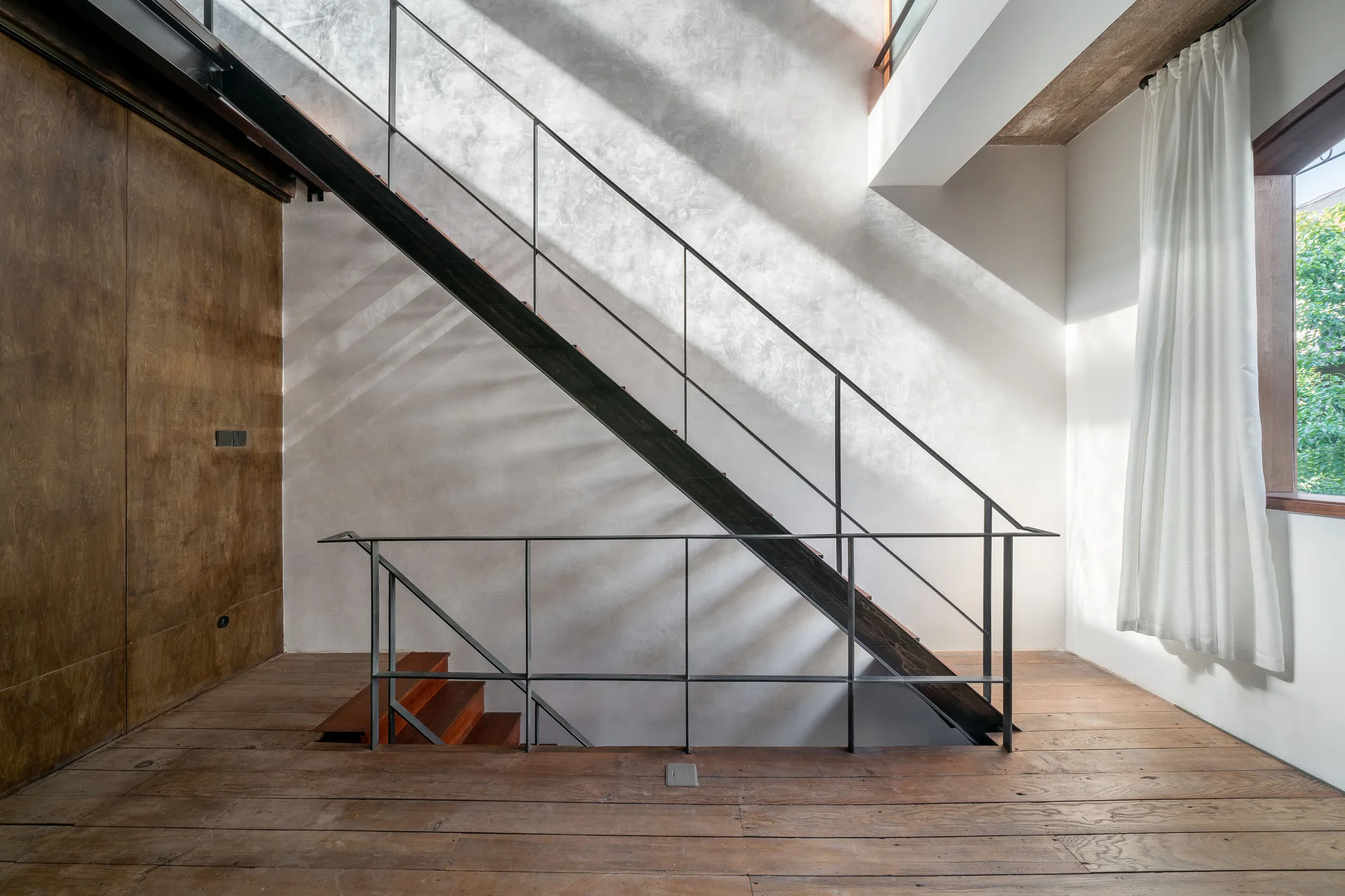 A minimalist staircase with a thin black metal railing stands against a textured grey plaster wall lit by dramatic natural sunlight.