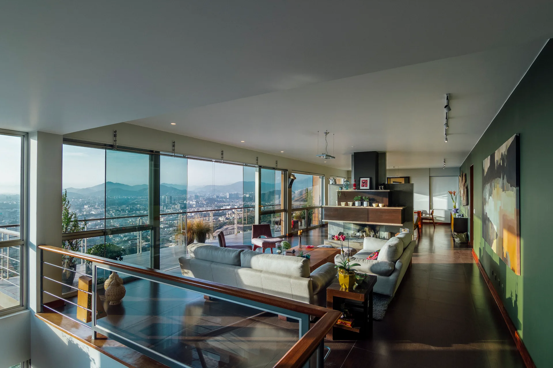 A contemporary living room featuring light gray sofas, a central fireplace, a deep green accent wall, and large glass windows with panoramic city views.