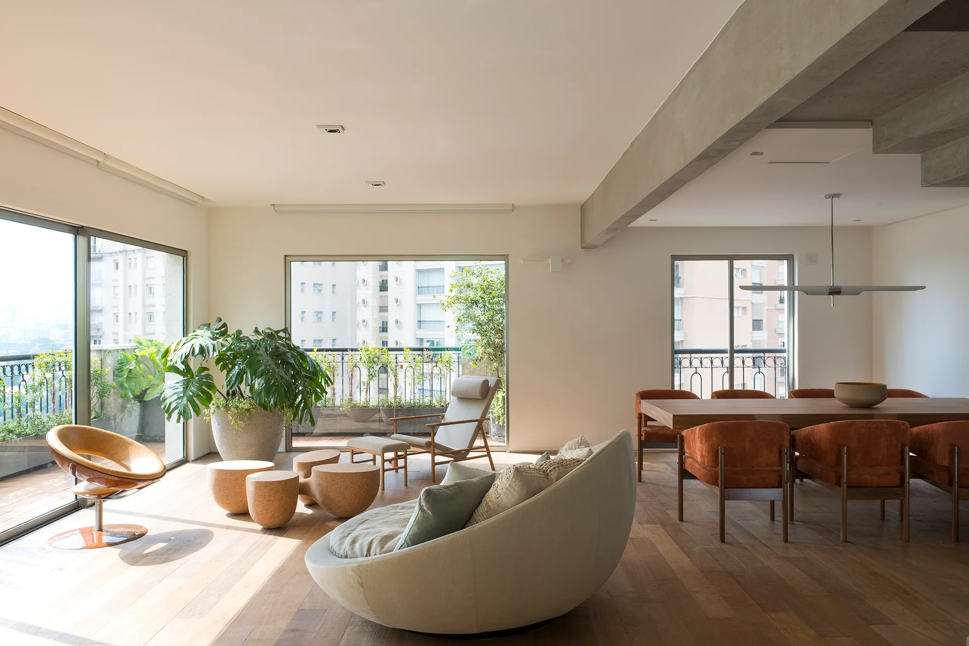 An open-plan living and dining room with a curved beige sofa, rust-orange dining chairs, wood floors, and an exposed concrete ceiling beam.