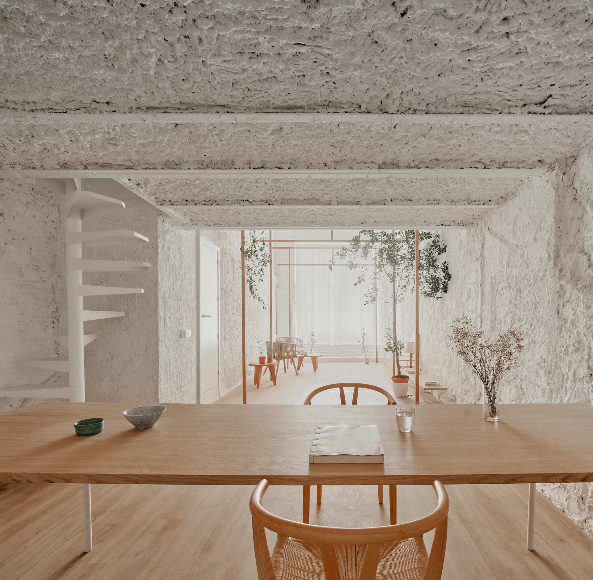 A minimalist dining area with a long light wood table, woven chairs, a white spiral staircase, and highly textured plaster walls and ceiling.