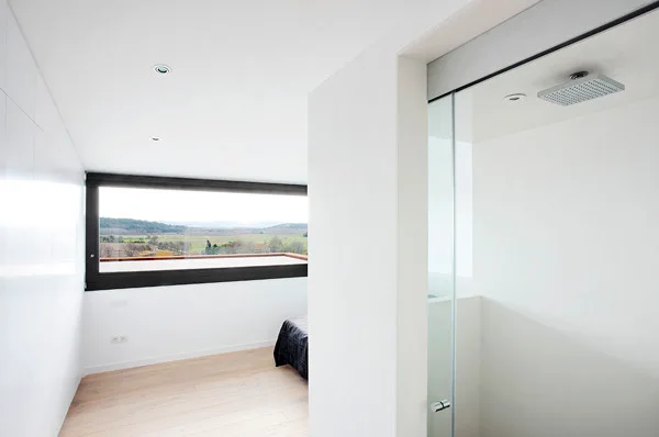 A minimalist white bedroom with light wood floors features a large panoramic window looking out to fields and a glass-enclosed shower in the foreground.