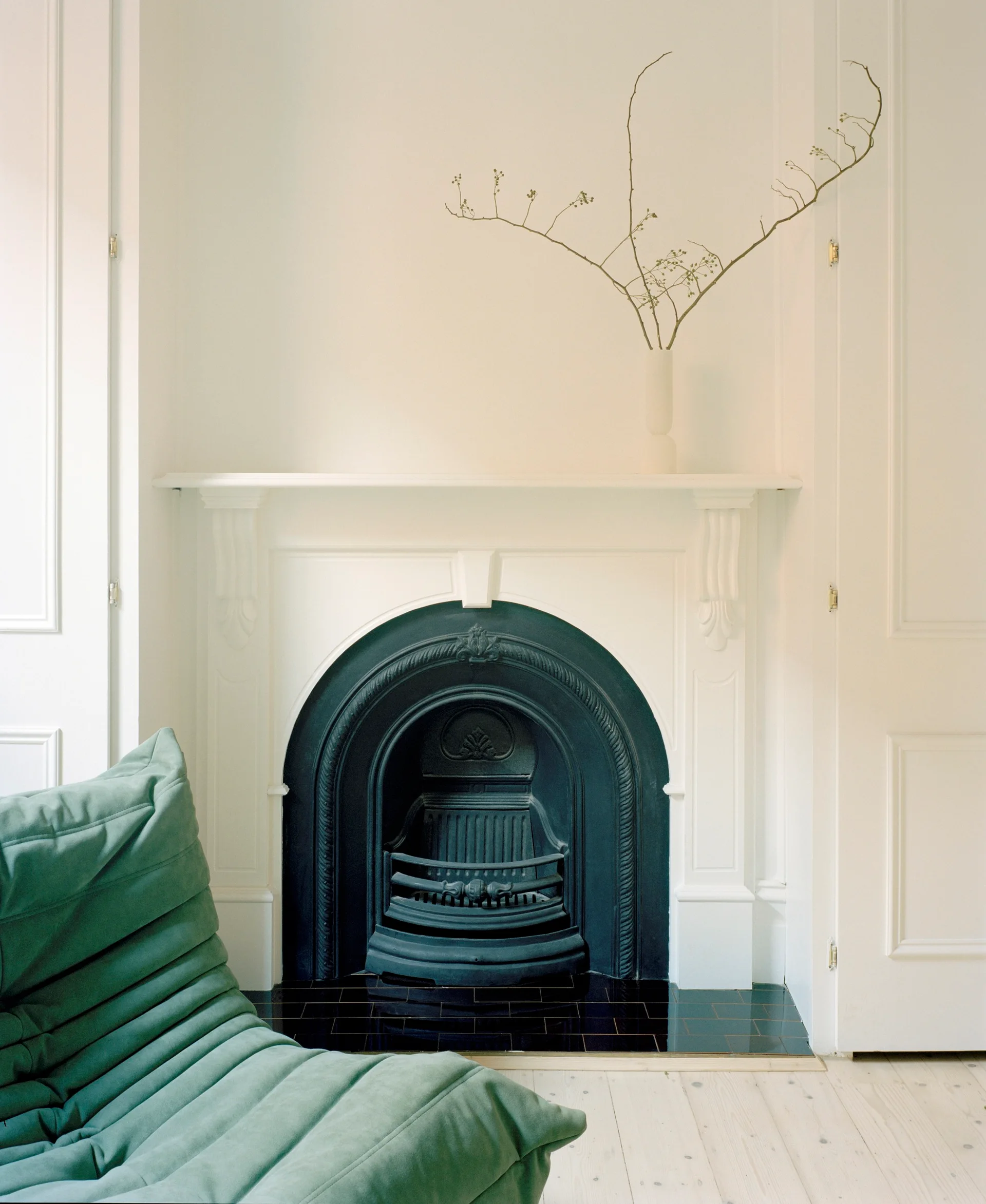 An arched black metal fireplace insert is framed by a white mantelpiece next to a green pleated armchair and white paneled doors.