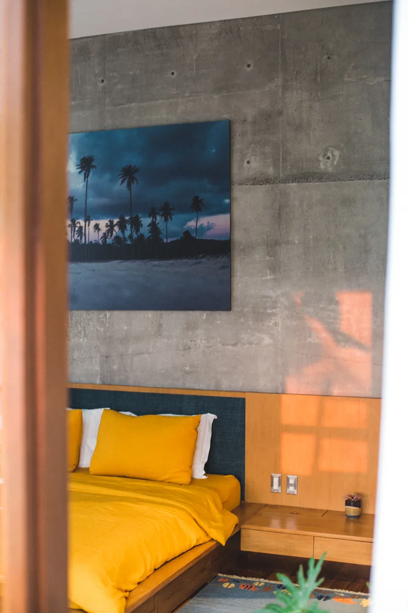 A modern bedroom features a bed with yellow bedding, an integrated wood headboard, and a raw concrete wall with a palm tree print.