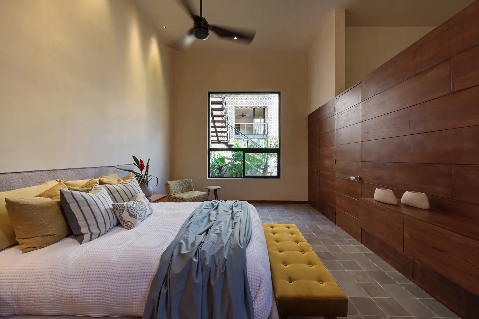 A modern bedroom with a large wood-paneled built-in closet wall, a bed with yellow and striped pillows, and a yellow tufted bench at the foot.