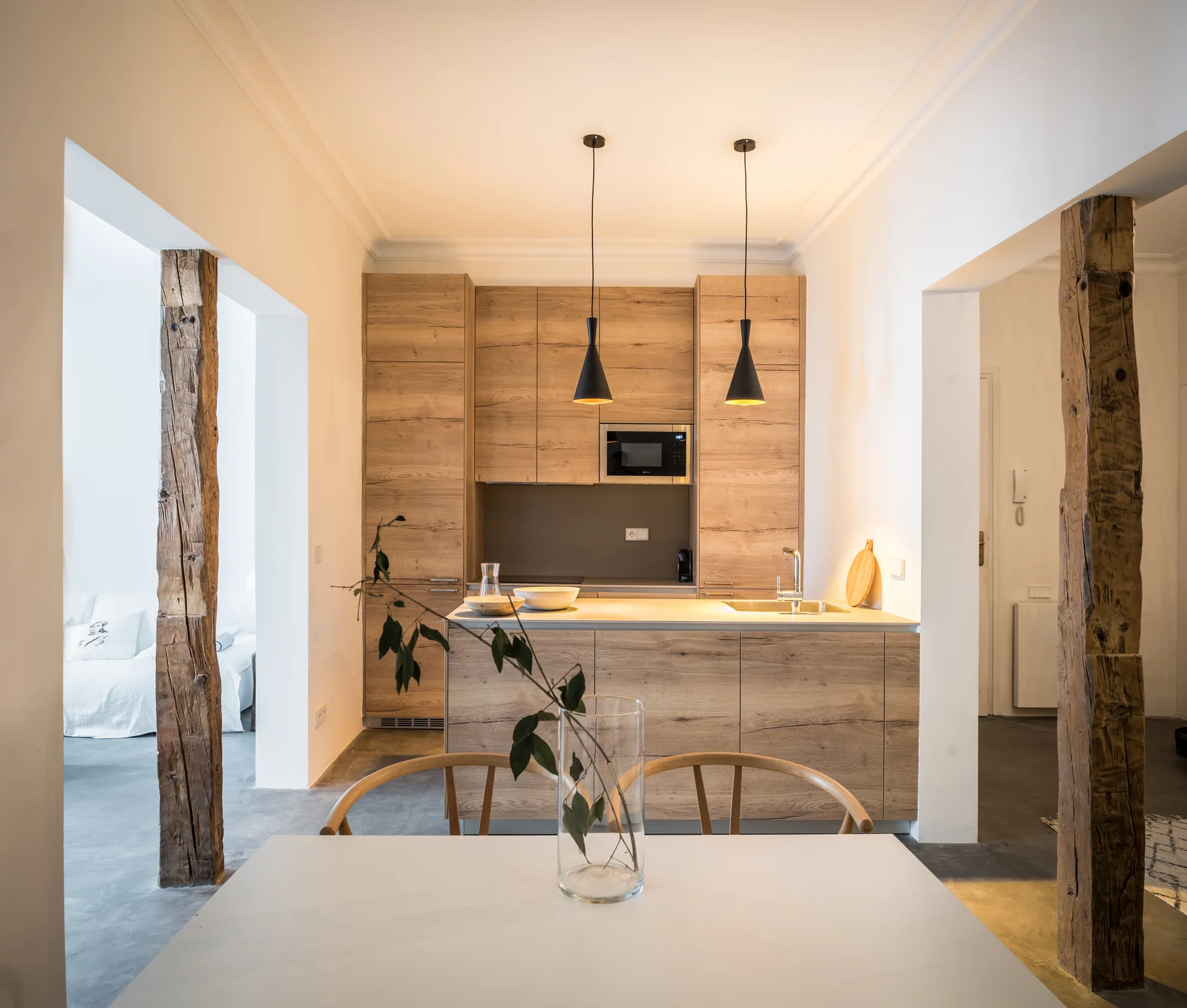 A modern wood-cabinetry kitchen and island are framed by two thick, rough-cut timber columns with a white dining table in the foreground.