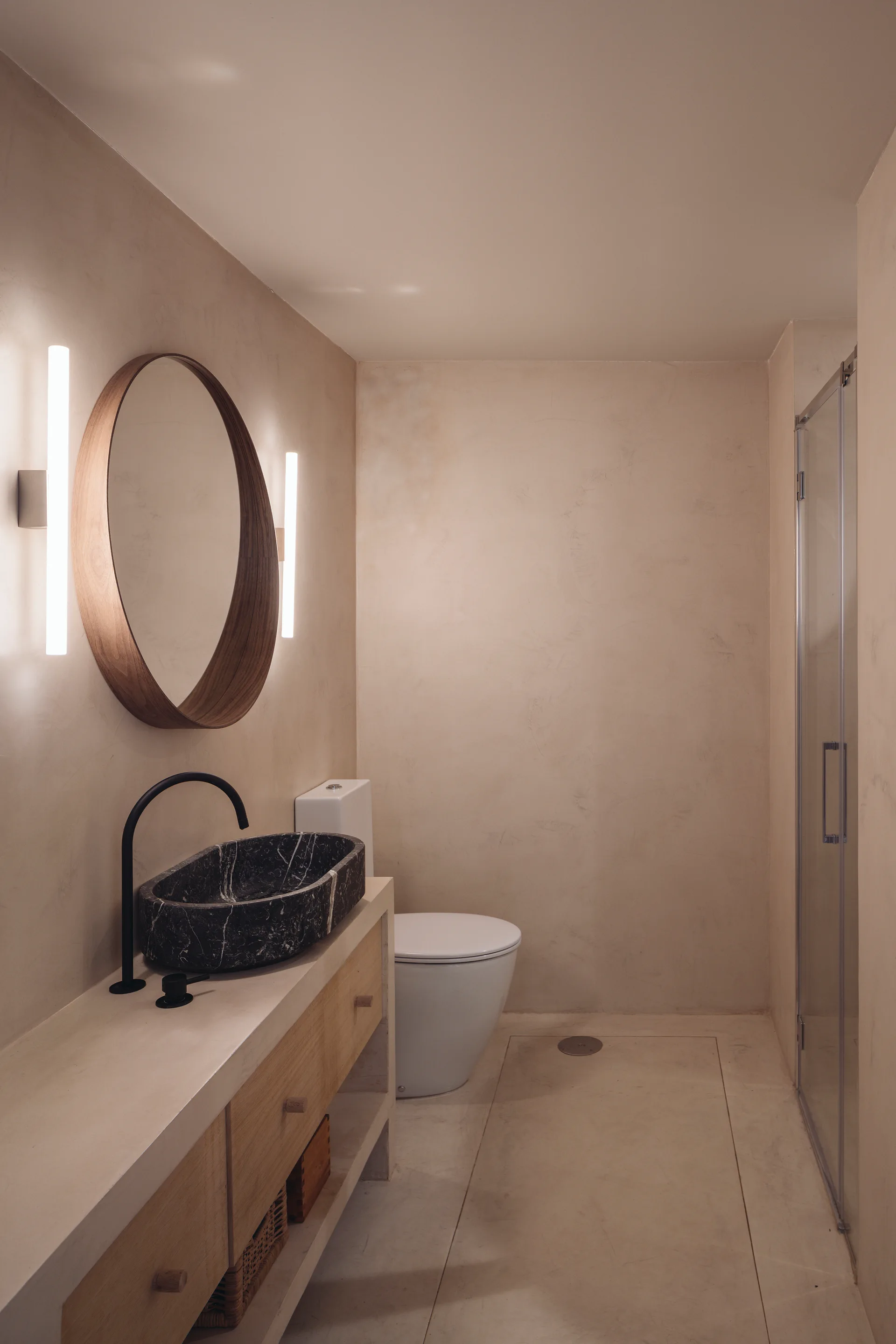 A minimalist bathroom features textured beige plaster walls, a round wood mirror with linear sconces, and a black stone vessel sink on a wood vanity.