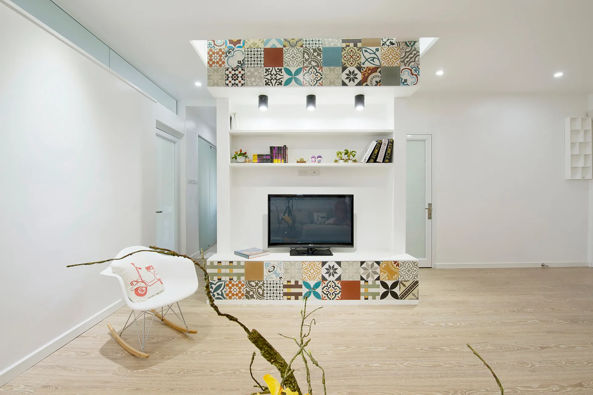 A white built-in media unit decorated with colorful patterned tiles houses a television and shelves beside a modern white rocking chair.