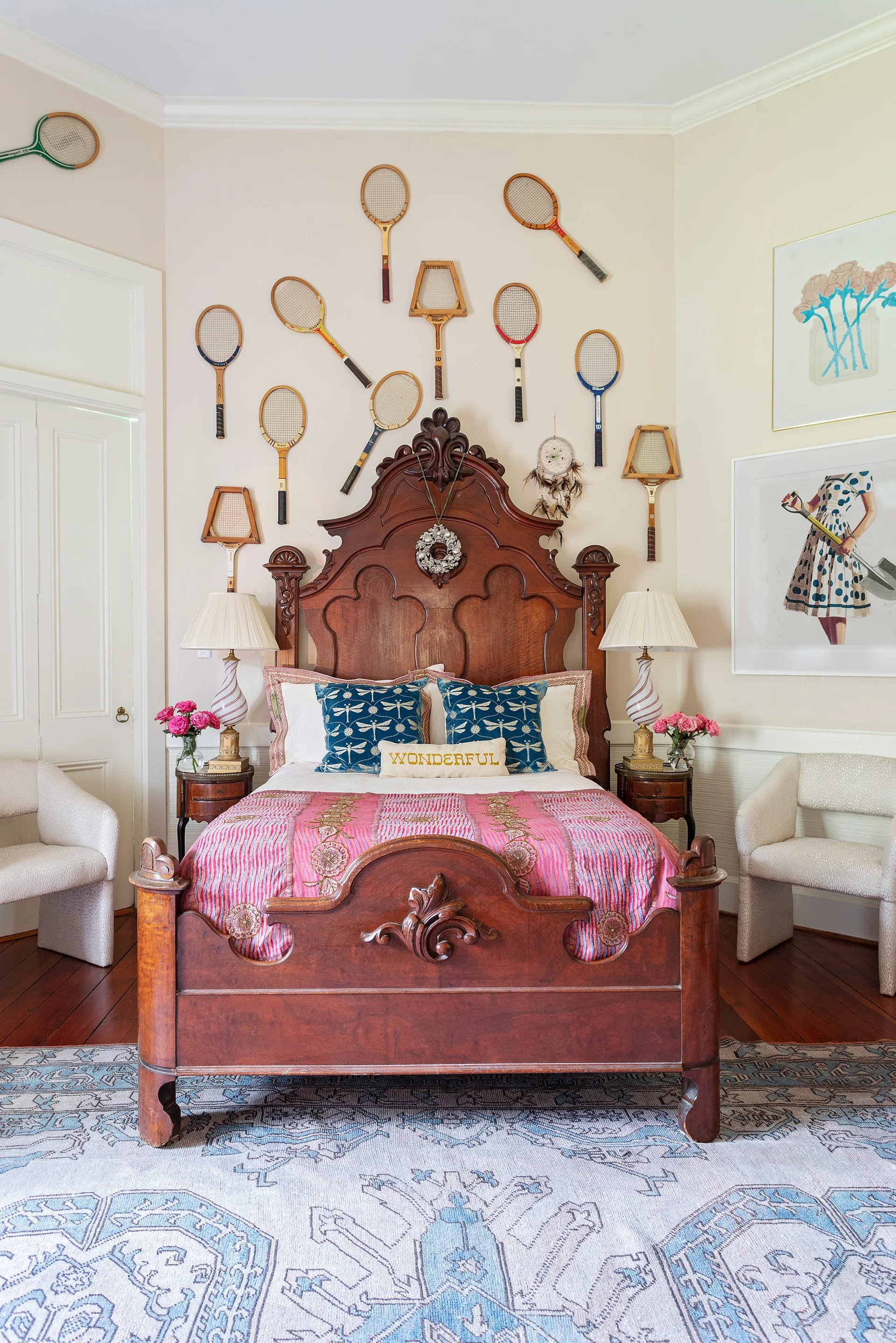 A bedroom featuring an ornate carved wooden bed frame, vintage tennis rackets mounted on the wall, two bedside lamps, and a patterned blue rug.
