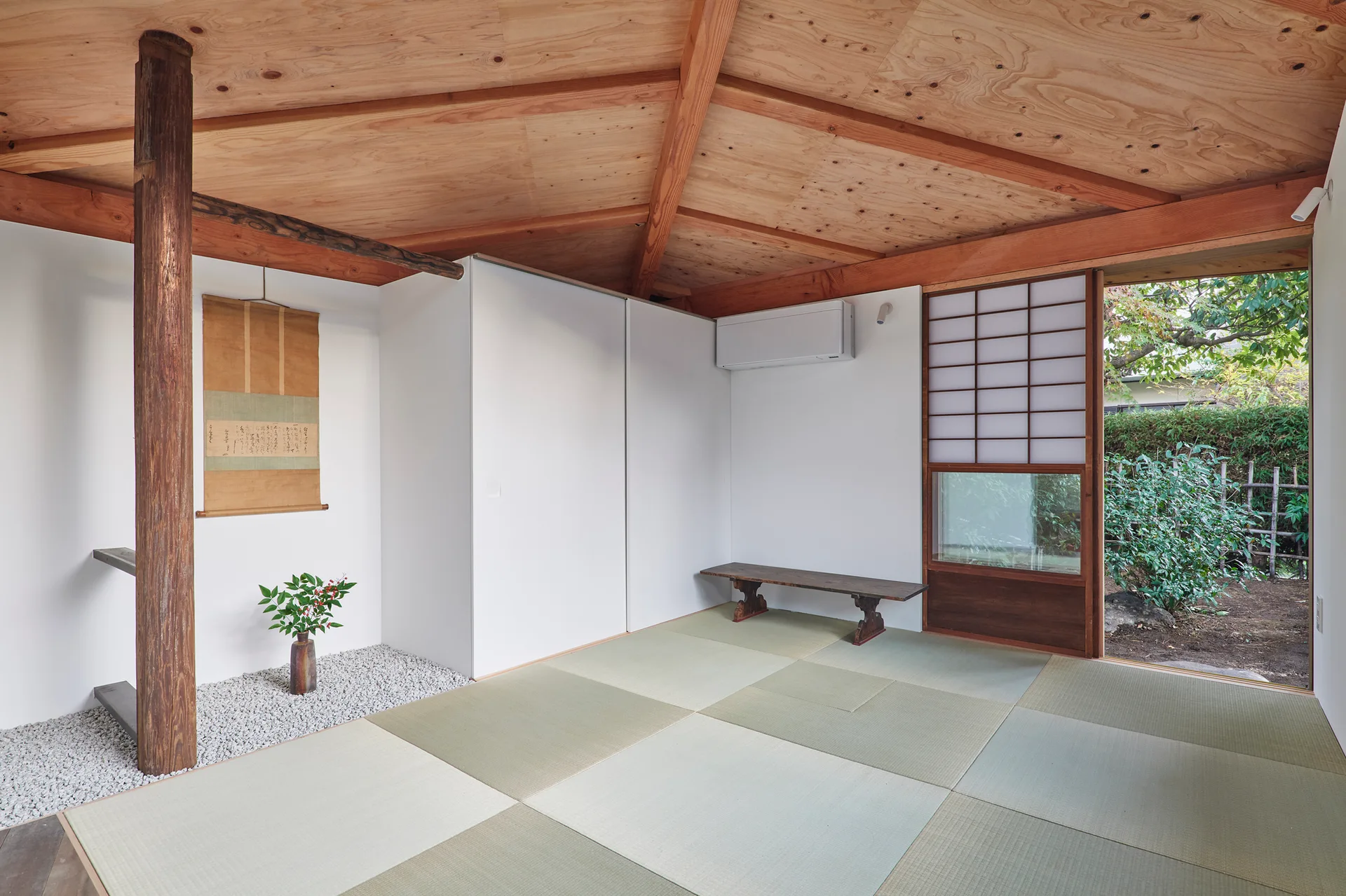 A minimalist Japanese-style room with green tatami mats, a wood-paneled ceiling, a shoji screen door, and a wooden pillar standing on white gravel.