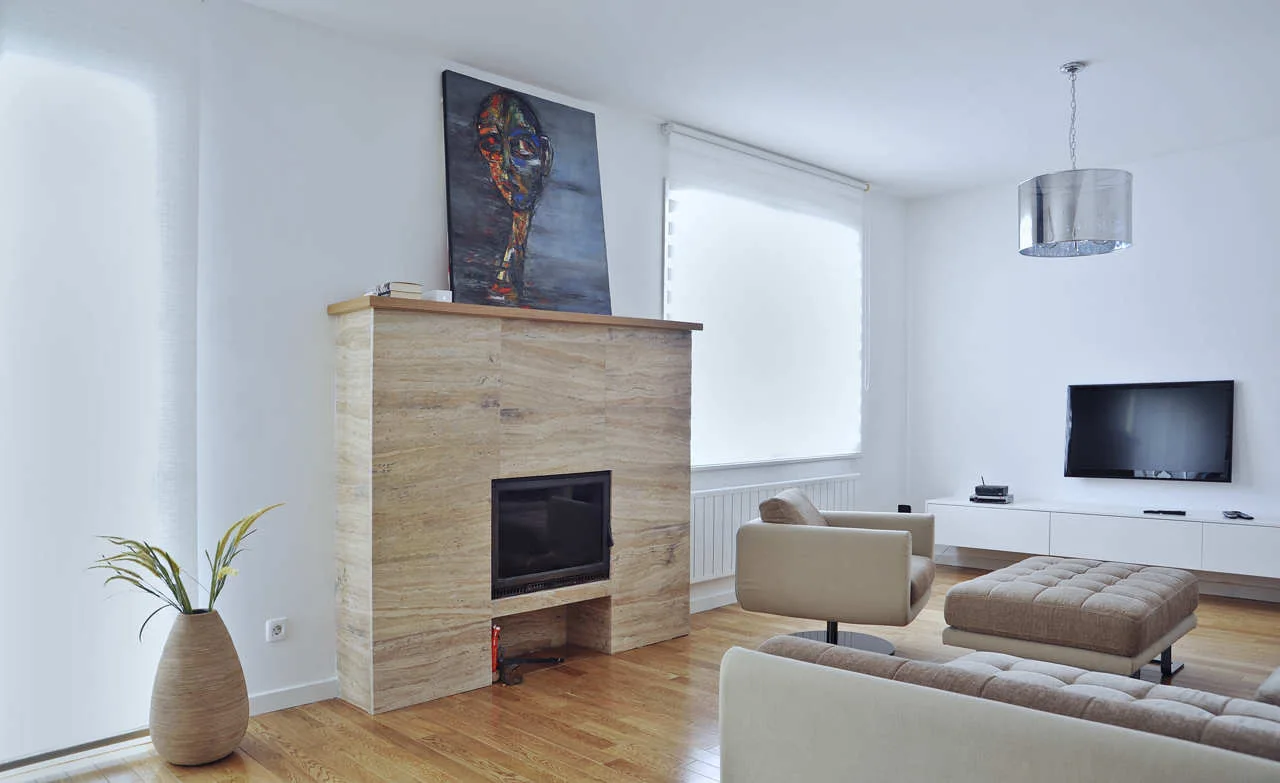 A minimalist living room featuring a tan stone fireplace, neutral tufted seating, a white media console, and a wall-mounted television.