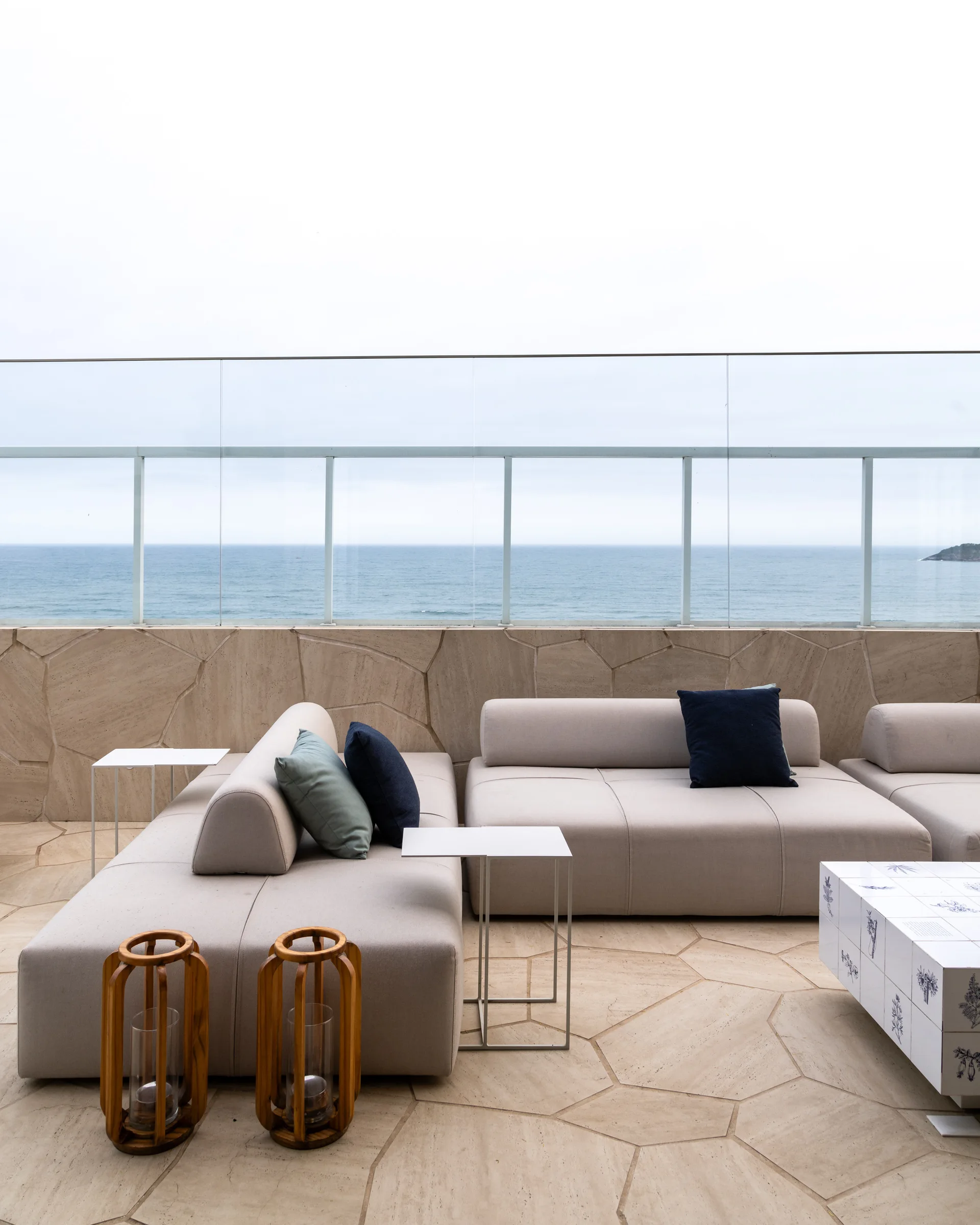A beige modular sectional sofa with blue pillows sits on a stone tiled terrace next to a glass railing overlooking the ocean.