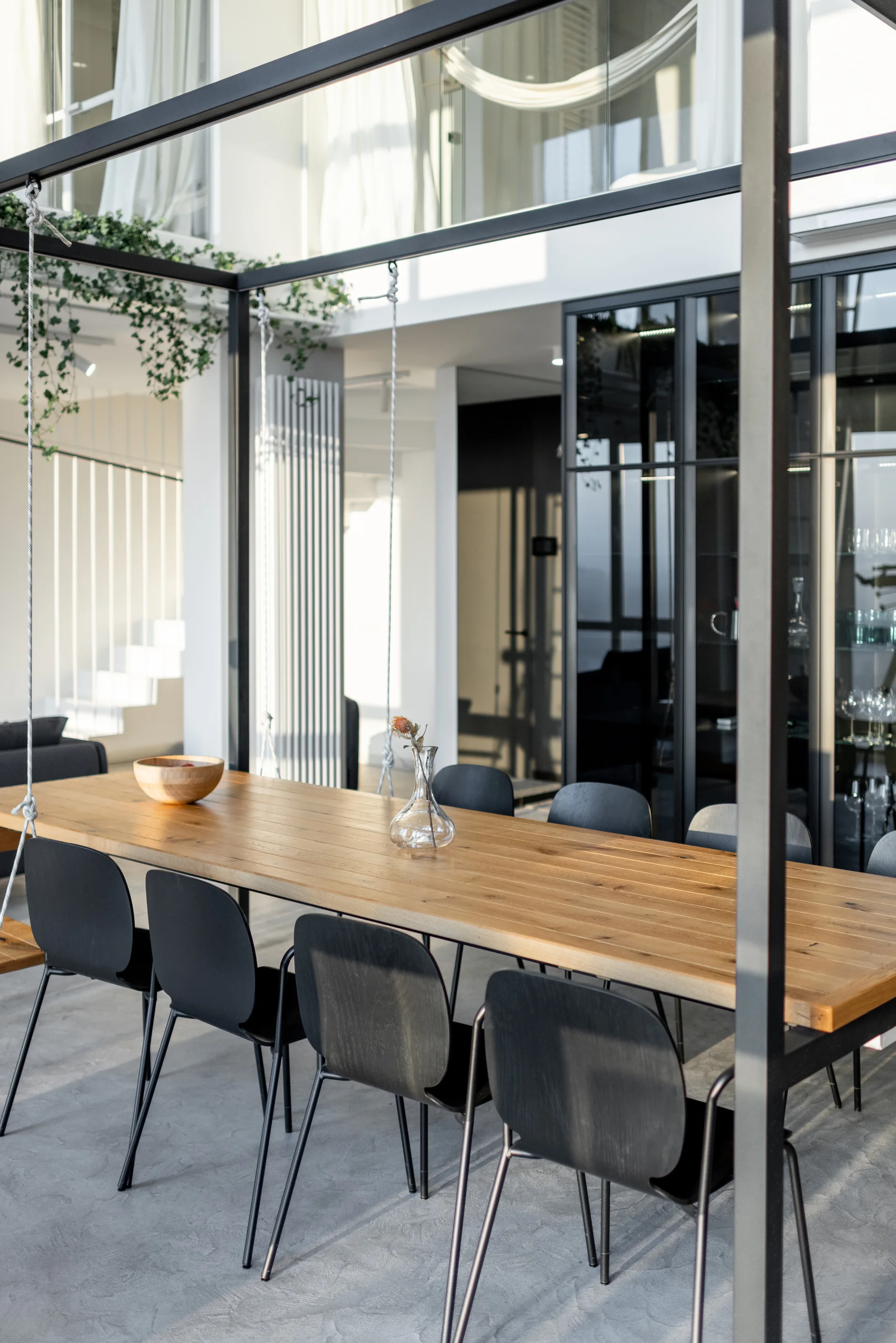 A long light wood dining table with black chairs sits on a concrete floor beneath an architectural black metal frame.