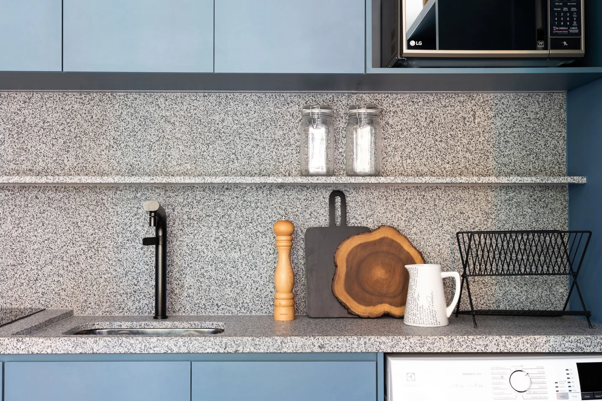 A kitchen counter with blue cabinets, a black faucet, and a speckled grey stone backsplash with an integrated open shelf.