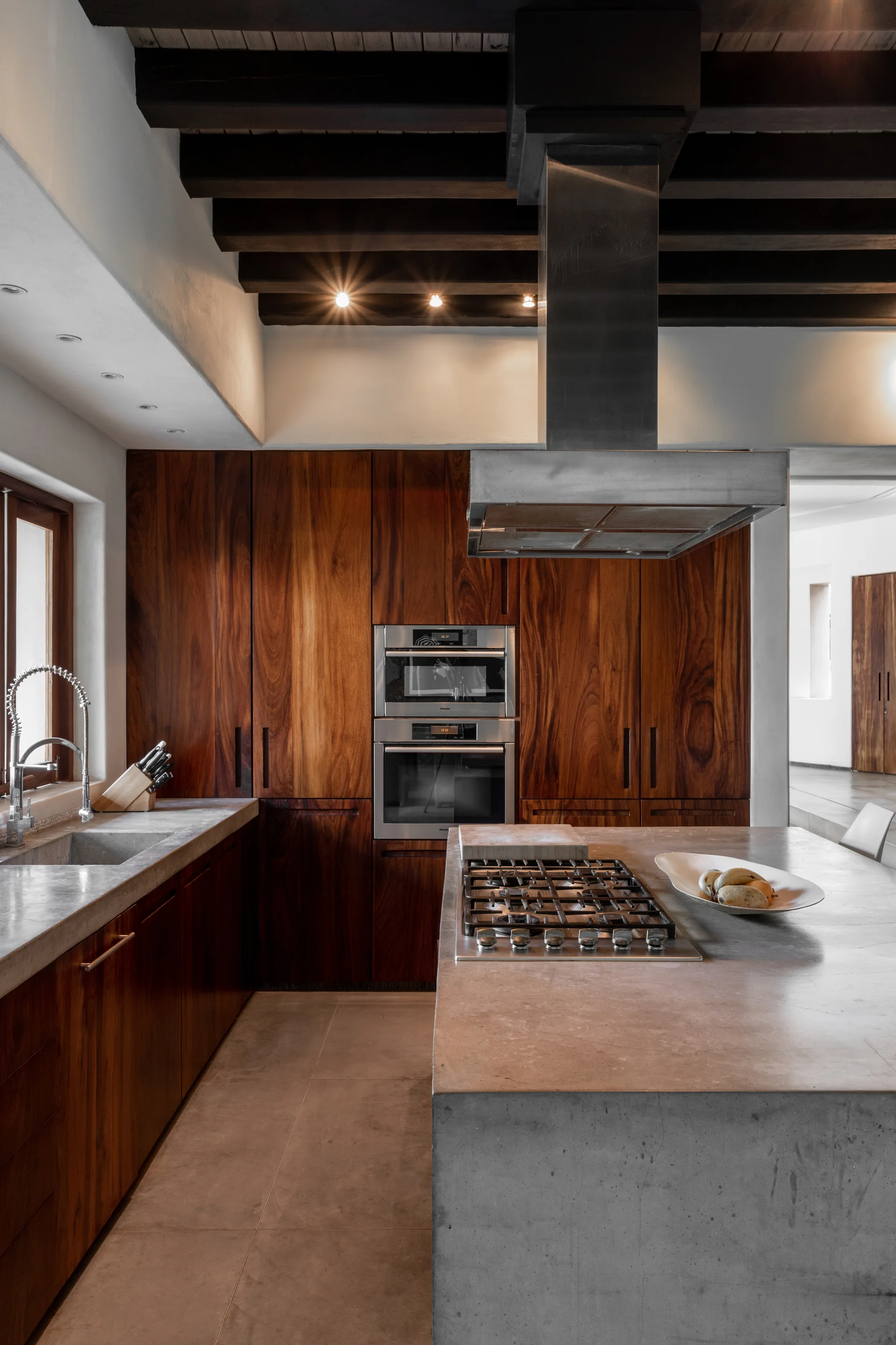 A modern kitchen features a poured concrete island with a gas range, dark wood cabinets, a built-in double oven, and exposed ceiling beams.
