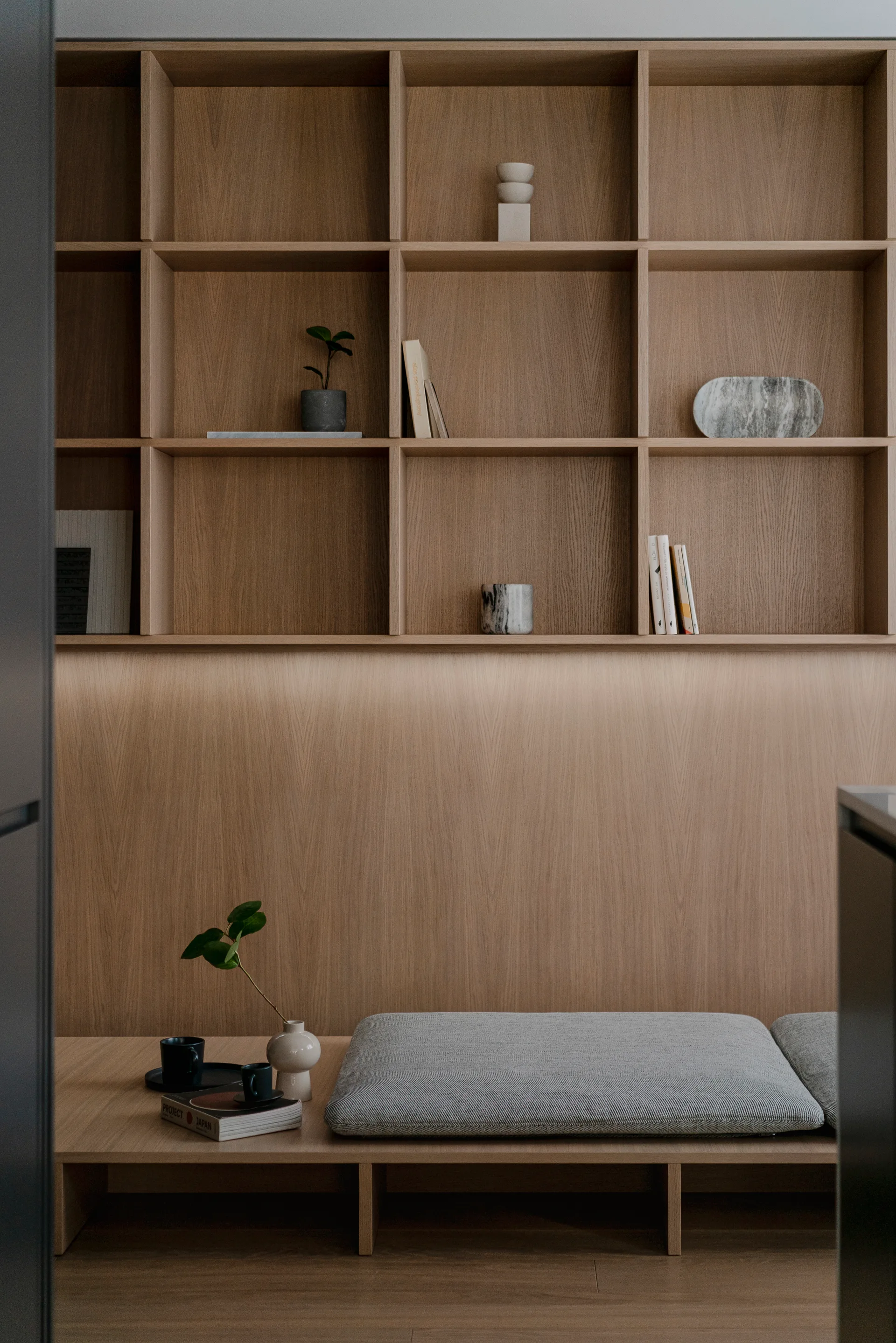 A custom light wood shelving unit with grid compartments stands above a low wooden bench featuring a grey fabric cushion and small decor items.