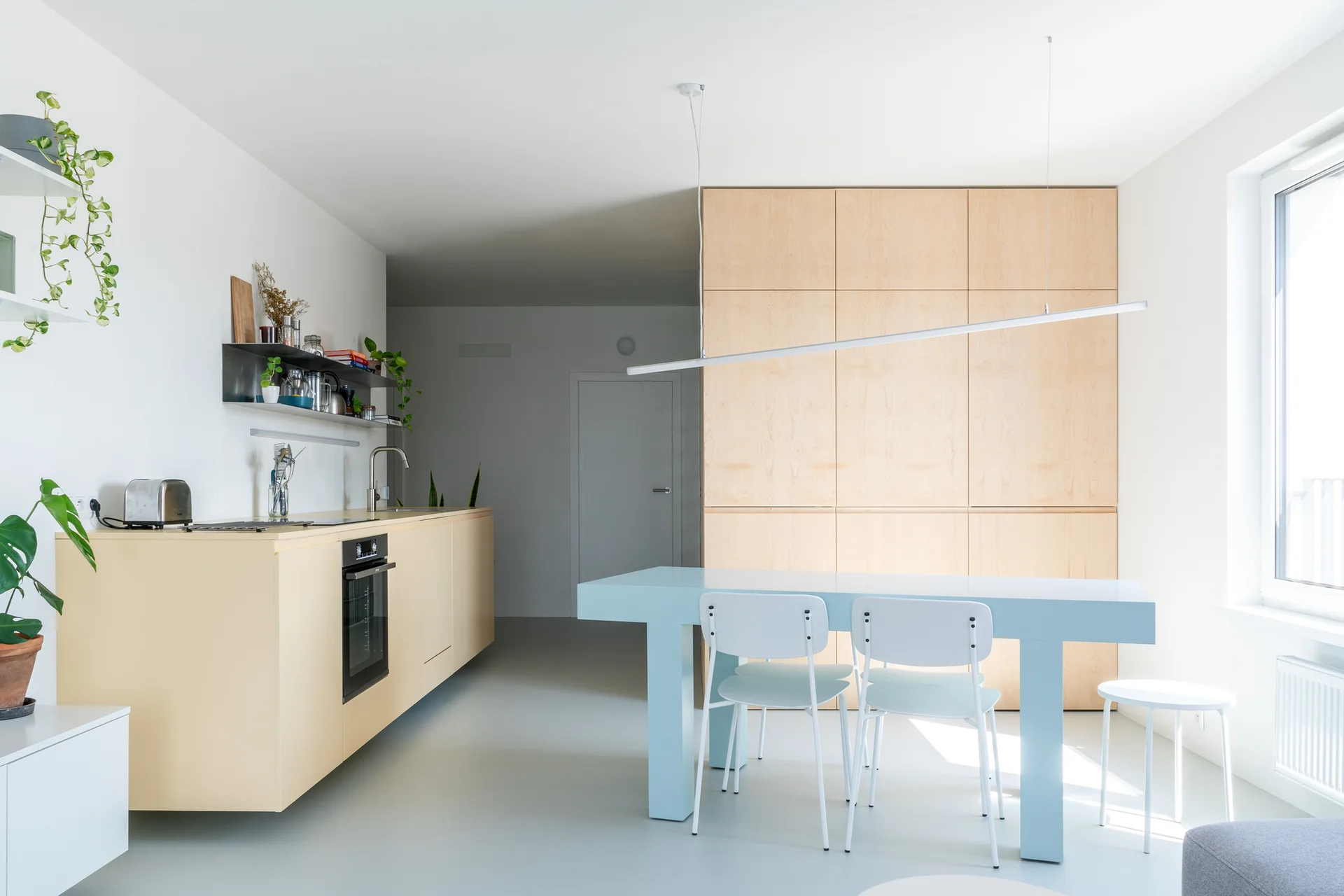 A minimalist kitchen features pale yellow cabinetry, a light blue dining table with white chairs, and a large light wood storage cabinet.