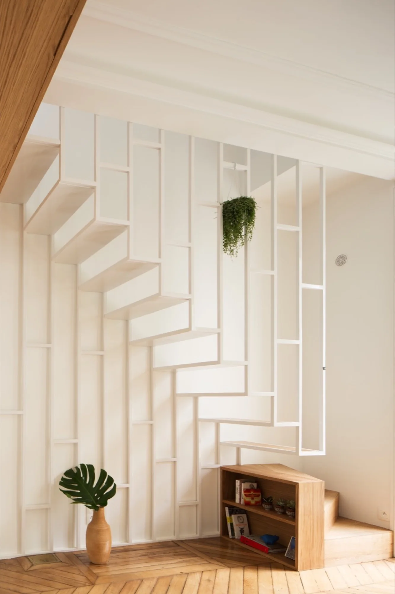 A modern staircase with a white geometric metal screen railing, light wood herringbone parquet floors, and a small wooden bookcase.