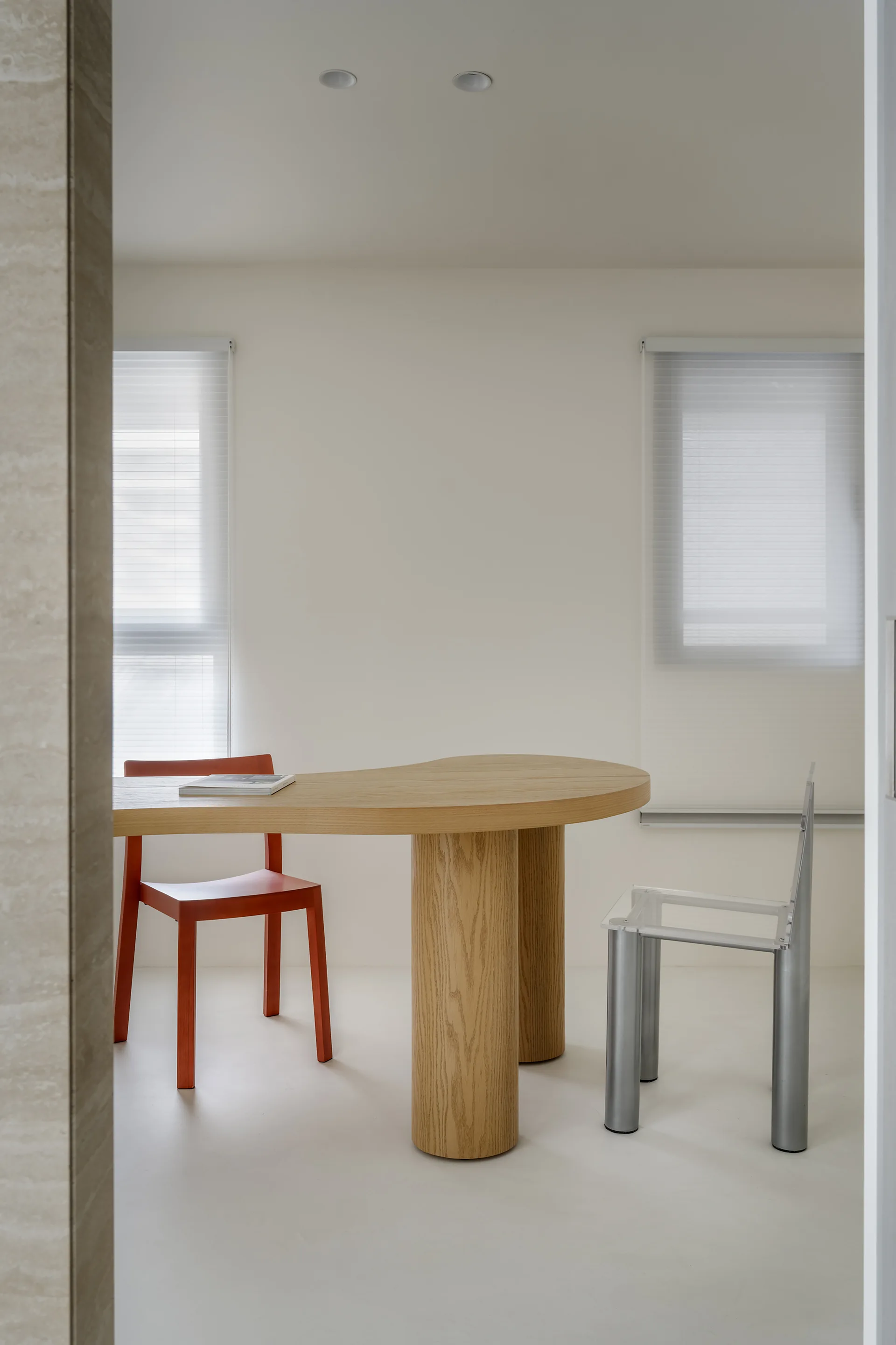 A minimalist dining room features a curved light wood table with a bright orange chair and a clear acrylic chair.