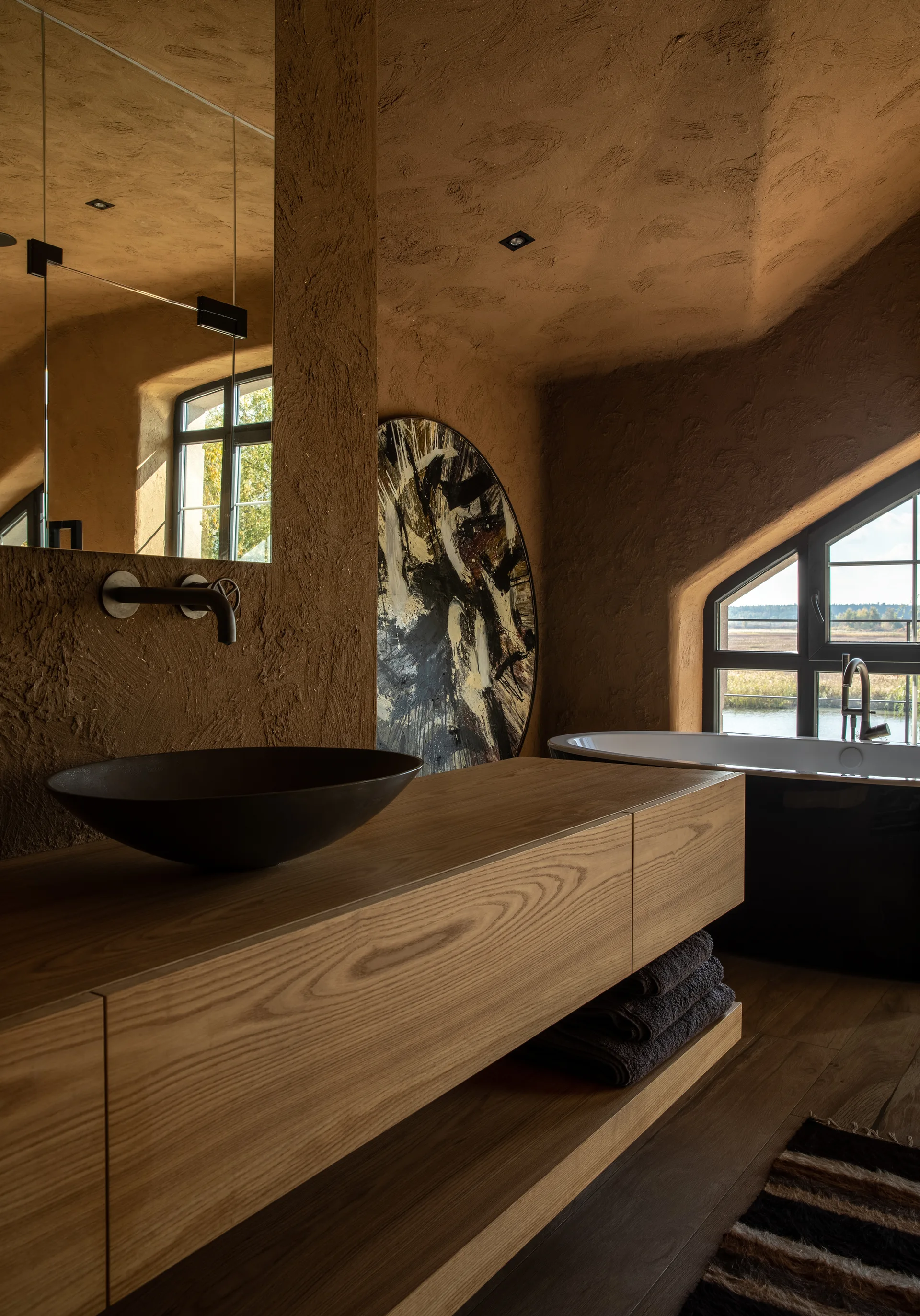 A modern bathroom features textured brown plaster walls, a floating wooden vanity with a dark bowl sink, and a black freestanding tub by a window.