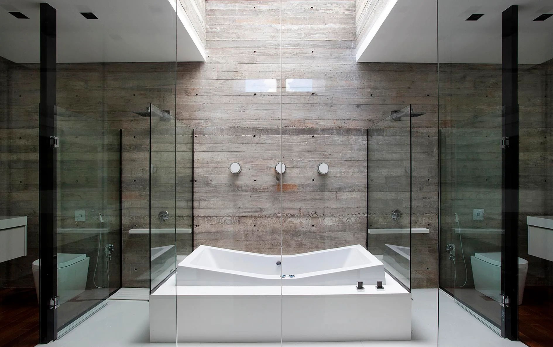 A geometric white bathtub stands centered beneath a bright skylight, surrounded by glass enclosures and a board-formed concrete wall.