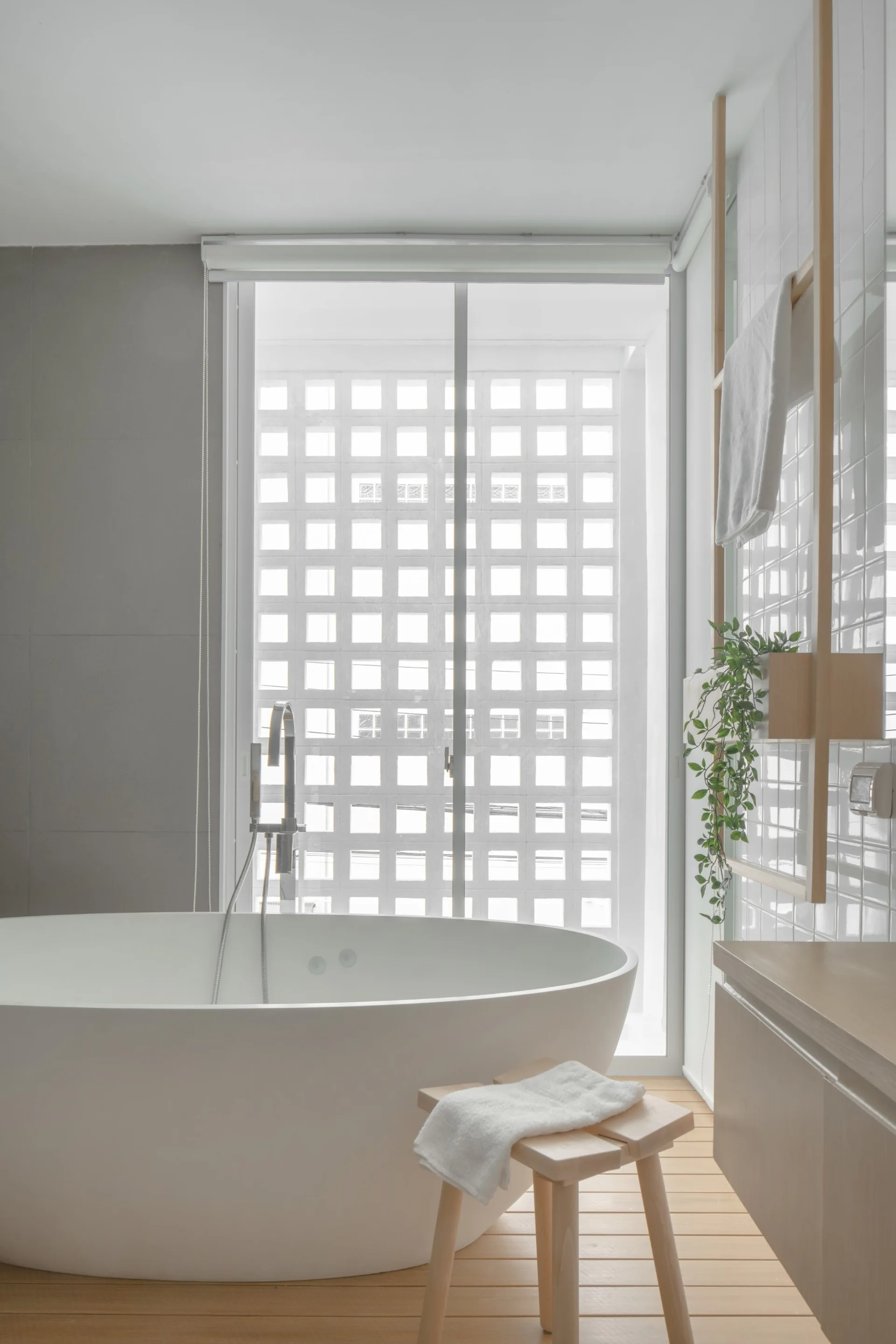 A minimalist bathroom features a white freestanding bathtub on a slatted wood floor in front of a window with an outdoor breeze block screen.
