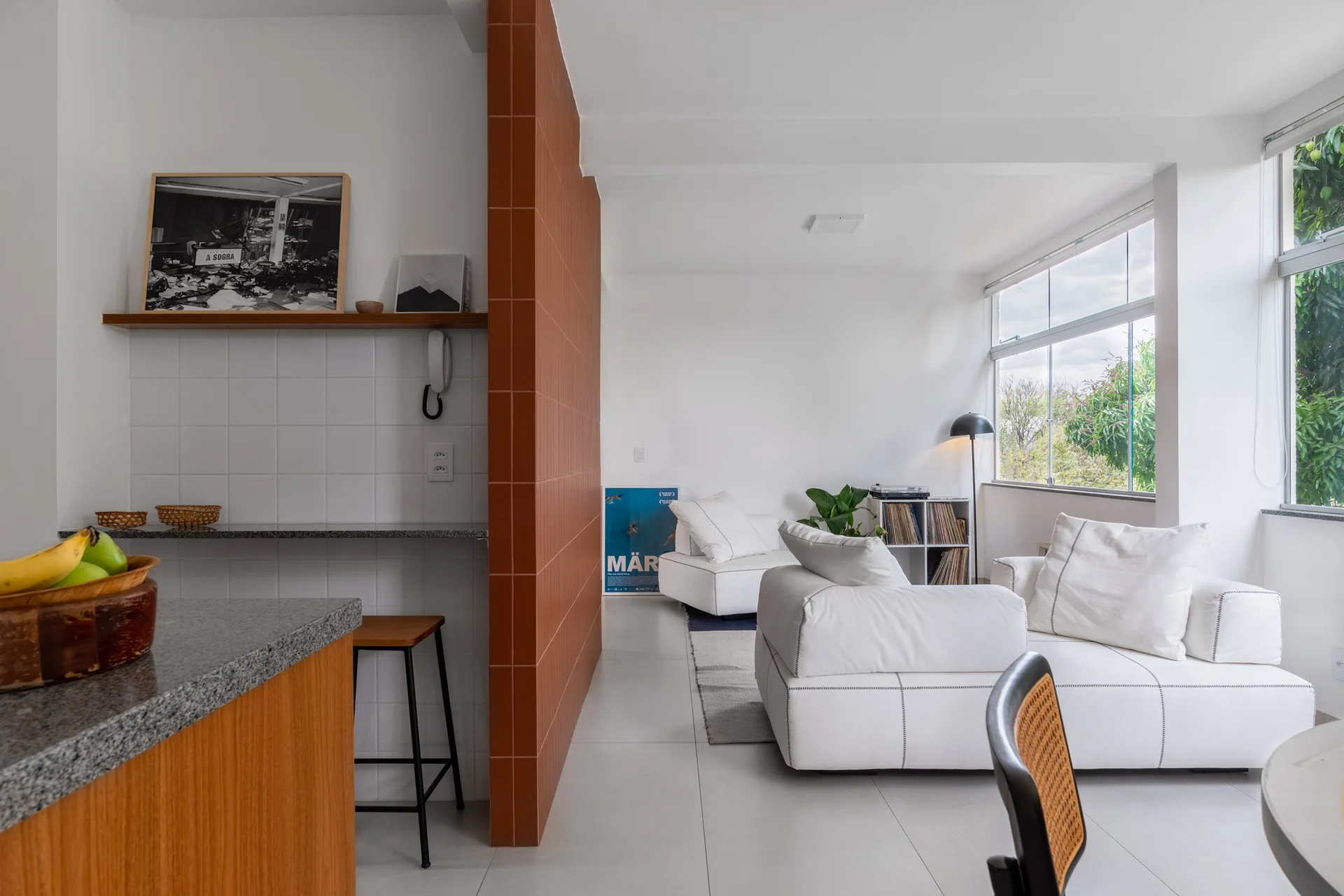 An open-plan apartment features a bright orange tiled partition wall separating a kitchen counter from a living room with a white modular sofa.