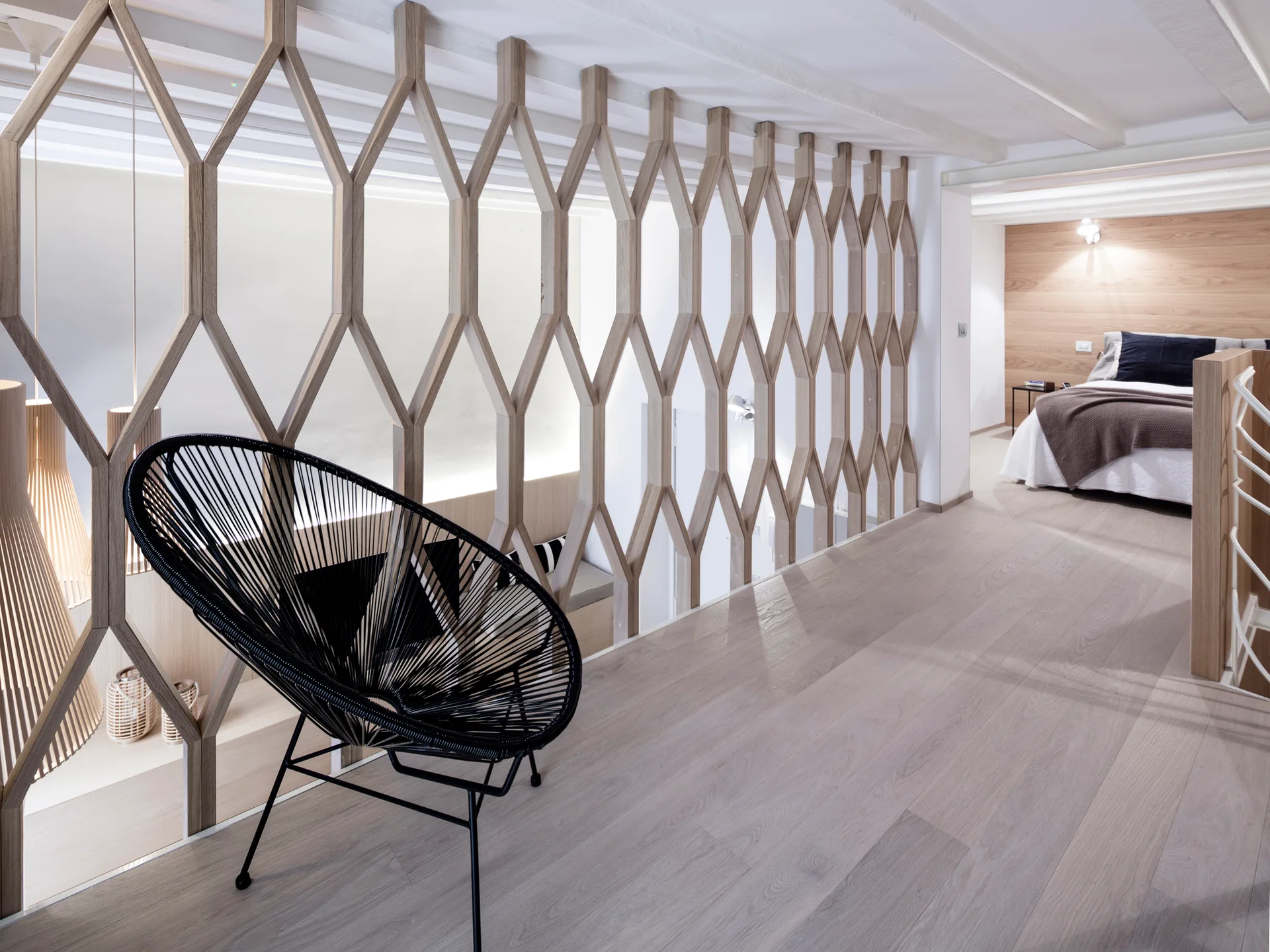 A modern bedroom hallway featuring a wooden diamond-lattice room divider, a black wire accent chair, and a bed in the background.