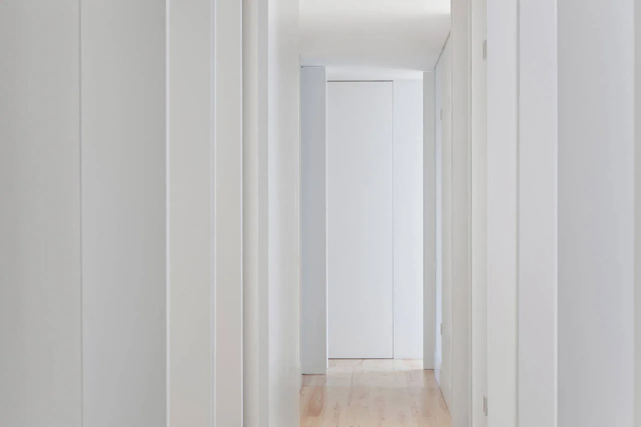 A minimalist white hallway features flush-mounted doors and light natural wood plank flooring.