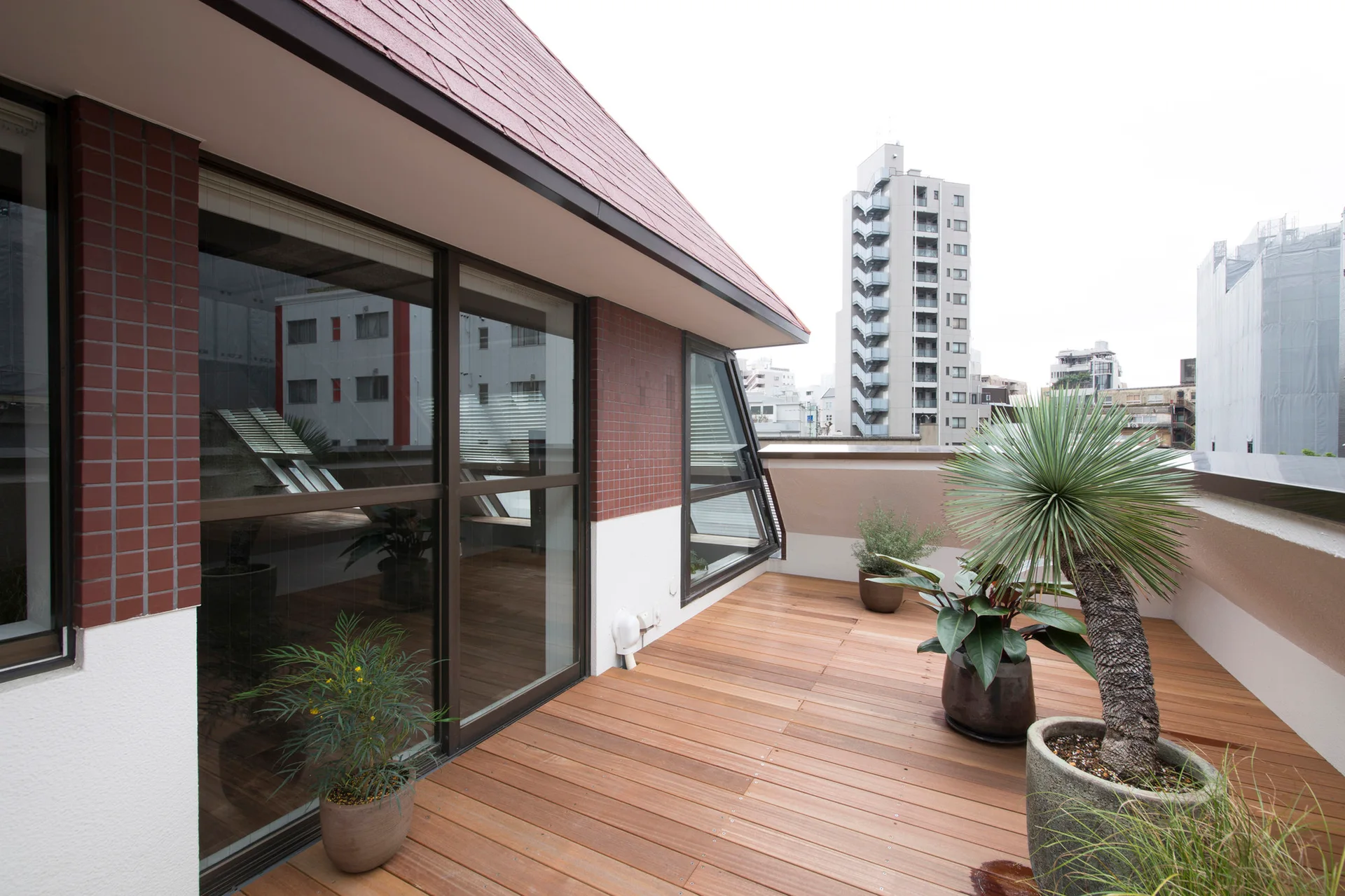 An urban rooftop terrace featuring a natural wood plank deck, sliding glass doors, red brick wall accents, and several potted green plants.
