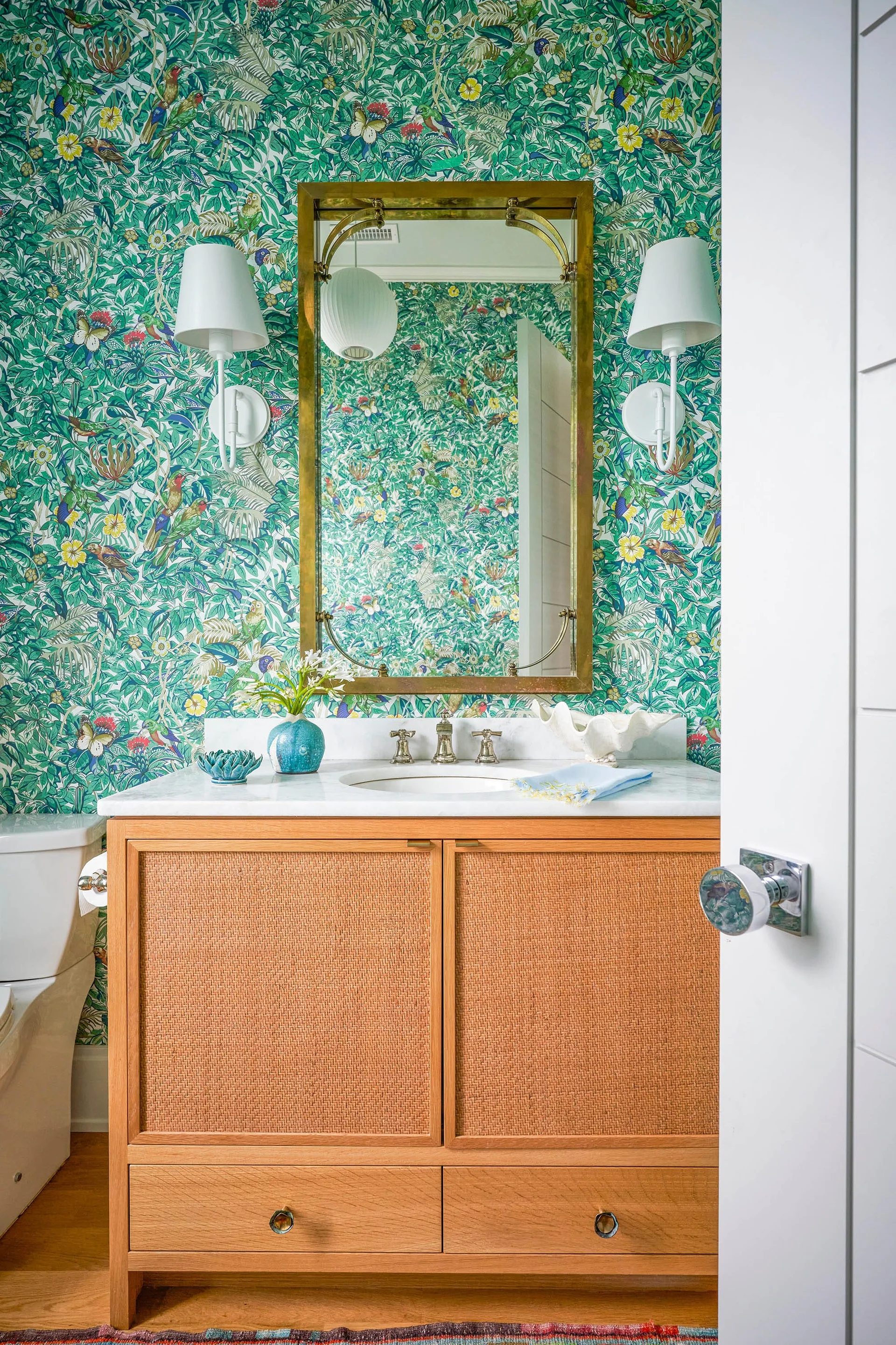A powder room features a natural wood vanity with rattan door panels, a white marble countertop, and vibrant green botanical wallpaper.