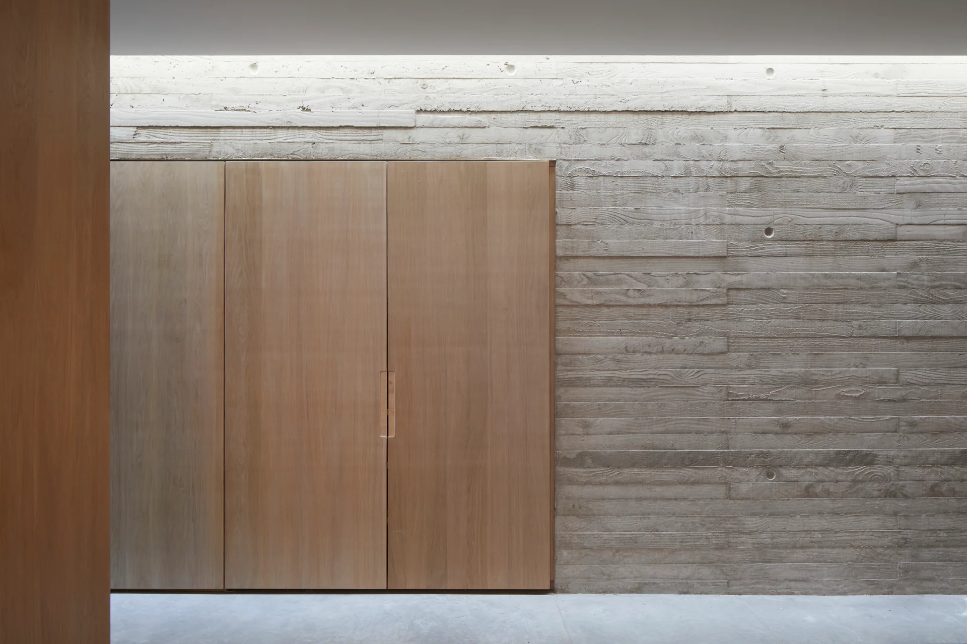 Flush-mount light wood cabinets built into a textured, board-formed concrete wall illuminated by a skylight.