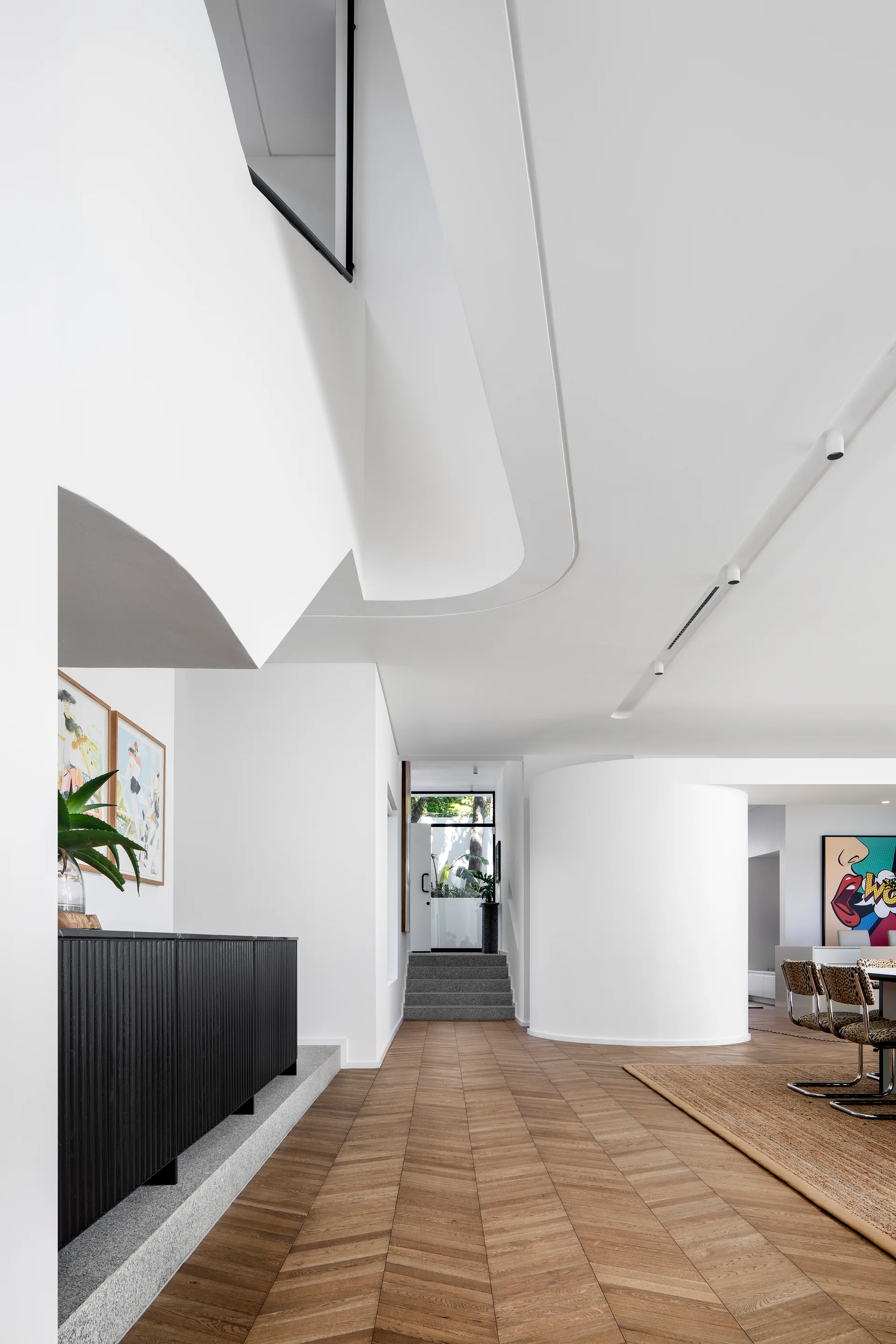 A modern entryway features white curved walls, chevron patterned wood flooring, a black ribbed sideboard, and stairs leading to a glass door.