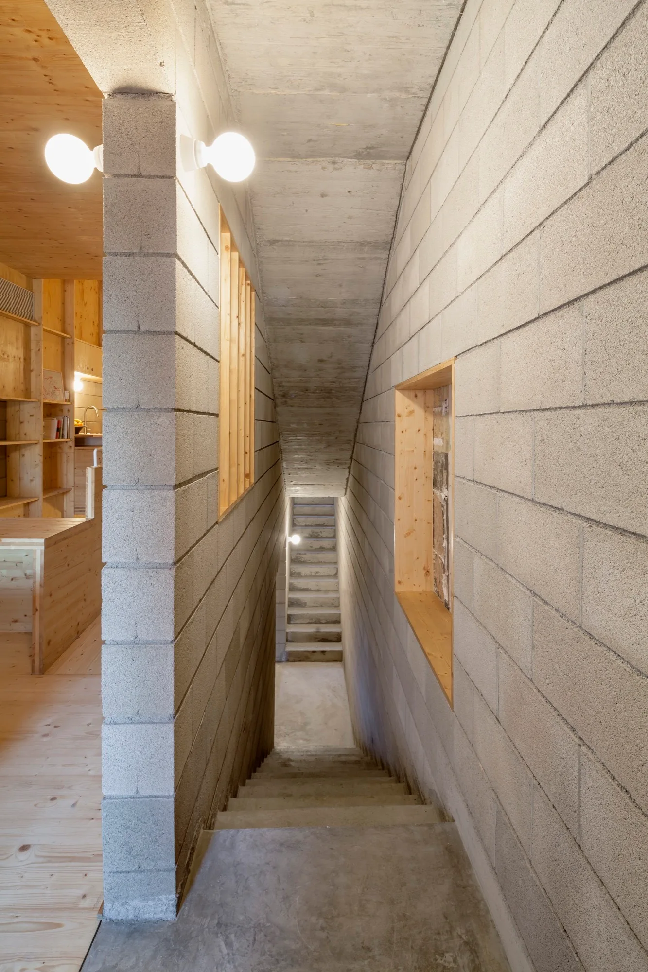 A narrow concrete staircase descends between light grey masonry block walls, with a wood-framed cutout looking into an adjacent pine-lined room.