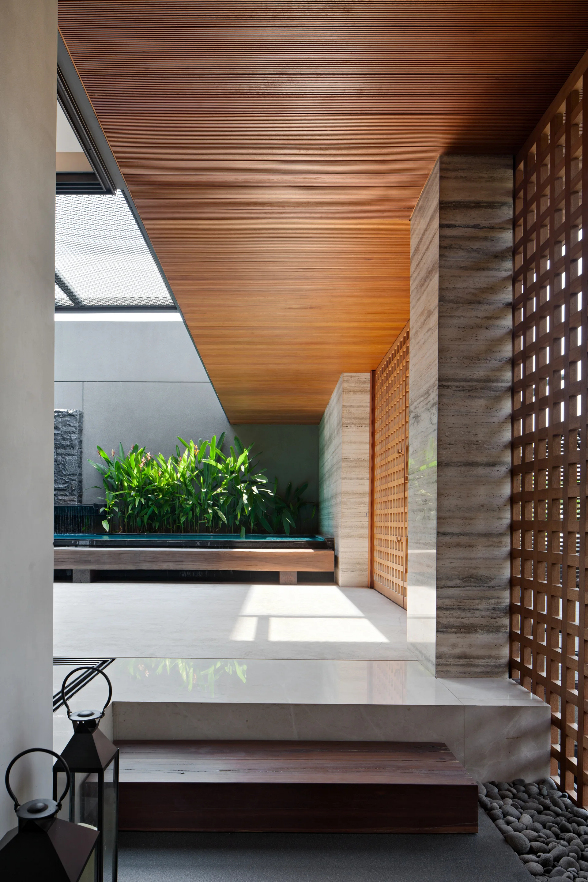 A modern covered patio features a wood slat ceiling, a textured stone pillar, a wooden lattice screen, and a water feature lined with green plants.