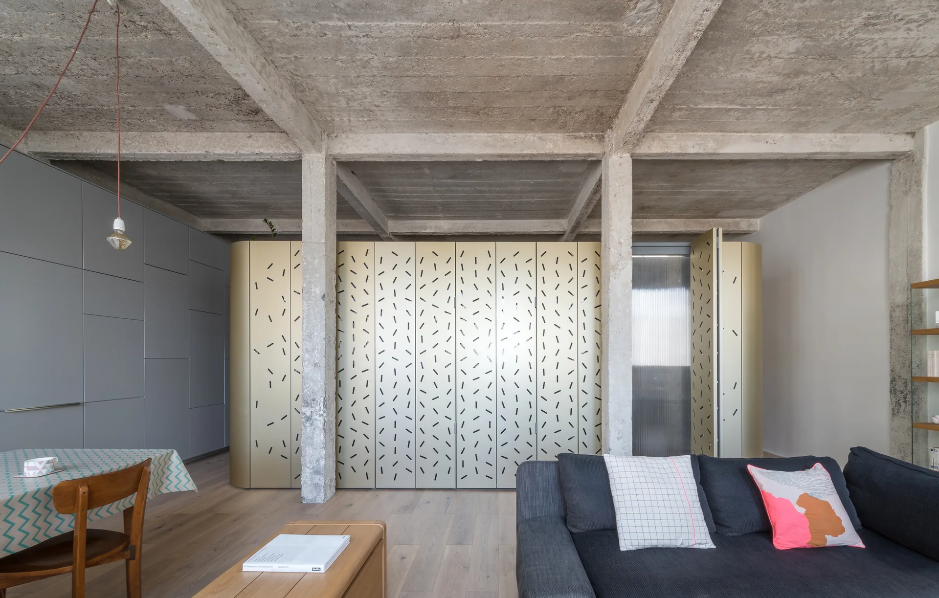 A modern living room features an exposed concrete ceiling, raw concrete columns, a charcoal sofa with pillows, and a large patterned gold storage cabinet.