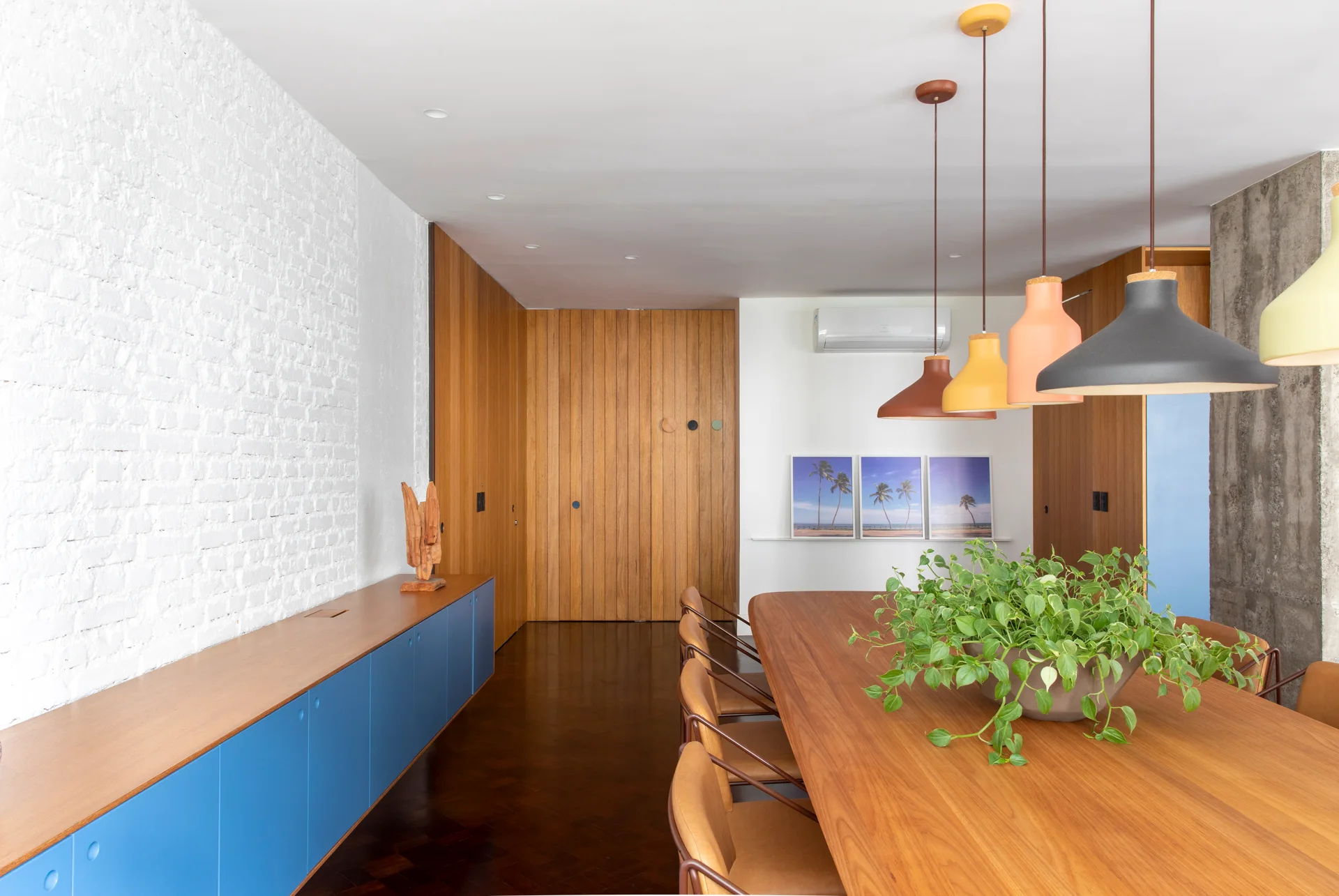A modern dining room featuring a long wooden table under colorful pendant lights, next to a blue cabinet against a white painted brick wall.