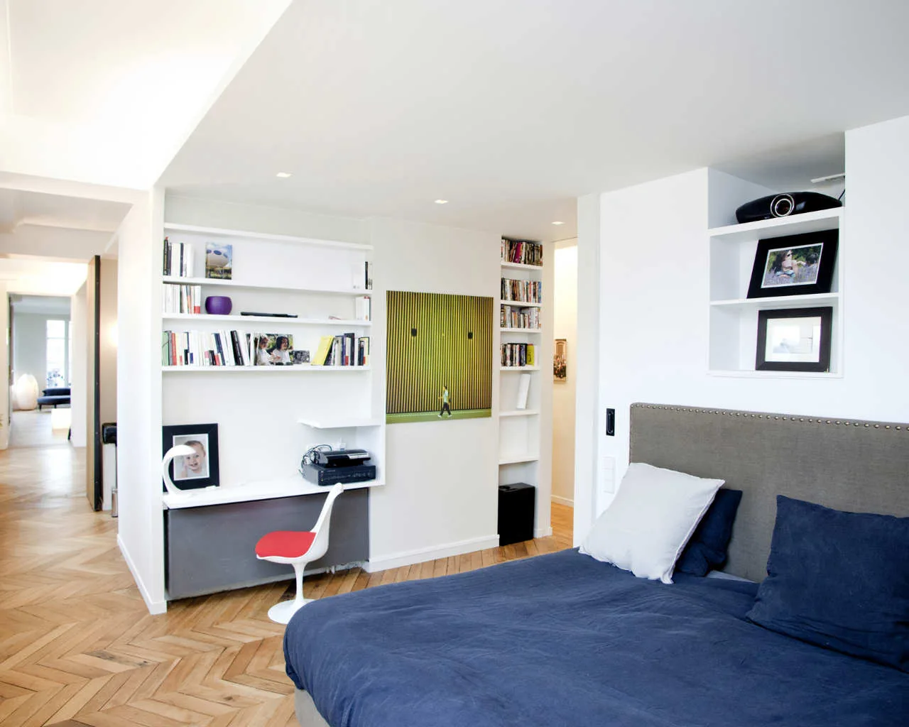 A modern bedroom featuring a bed with blue bedding, white built-in shelving with an integrated desk nook, and light-colored herringbone wood flooring.